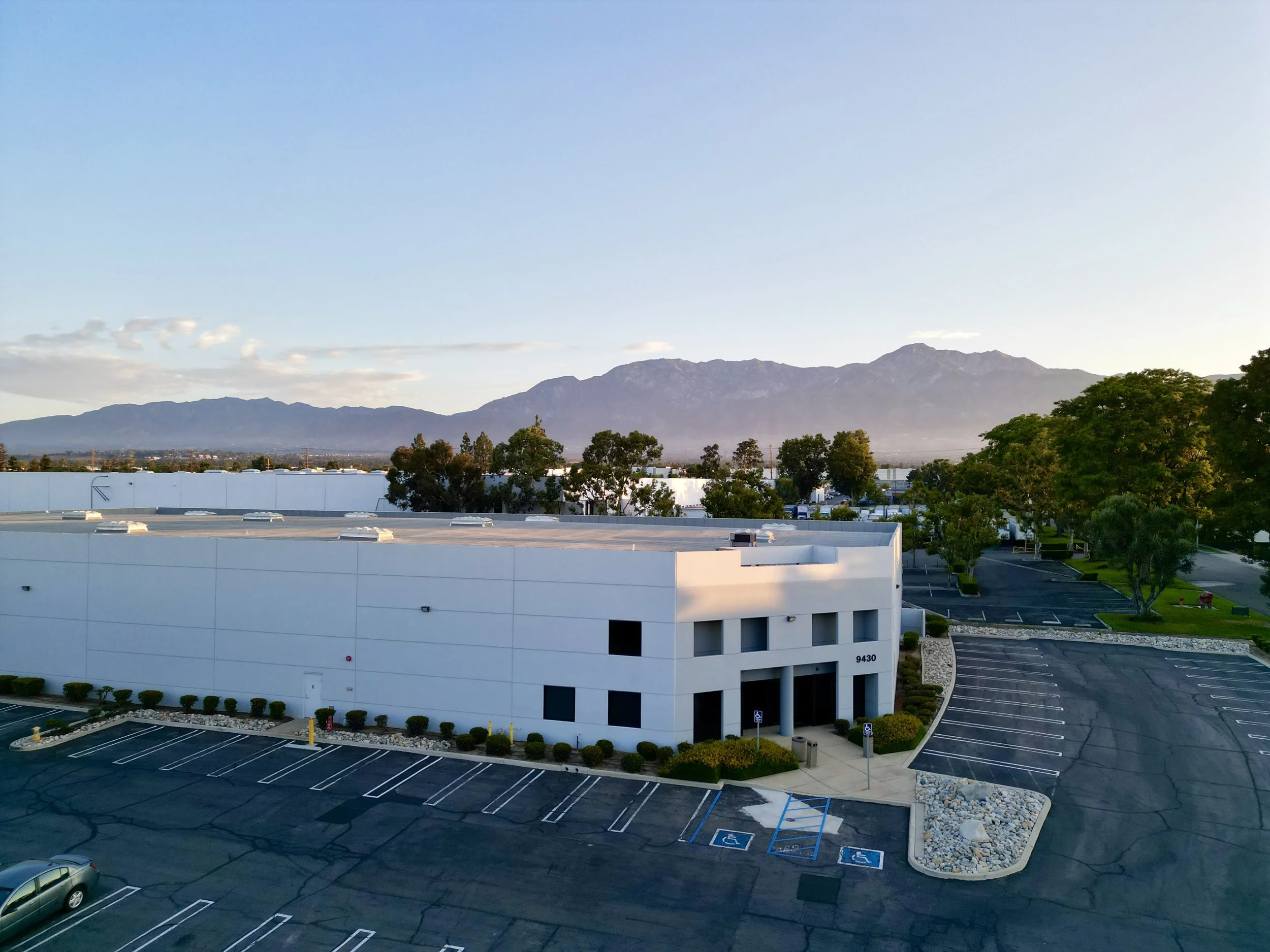 Empty parking lot in front of a white industrial warehouse building with mountains in the background.