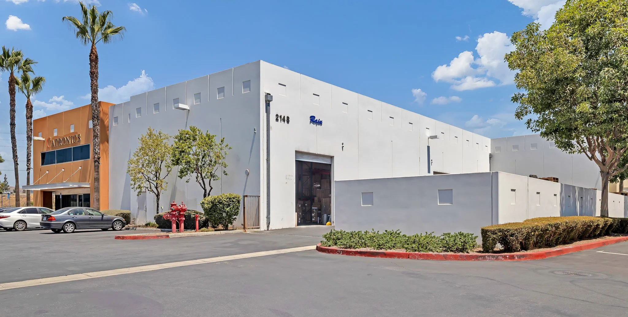A warehouse building with a sign reading 'CATRONICS,' a white facade, surrounded by a parking lot with cars, trees, and fire hydrants, under a partly cloudy sky.