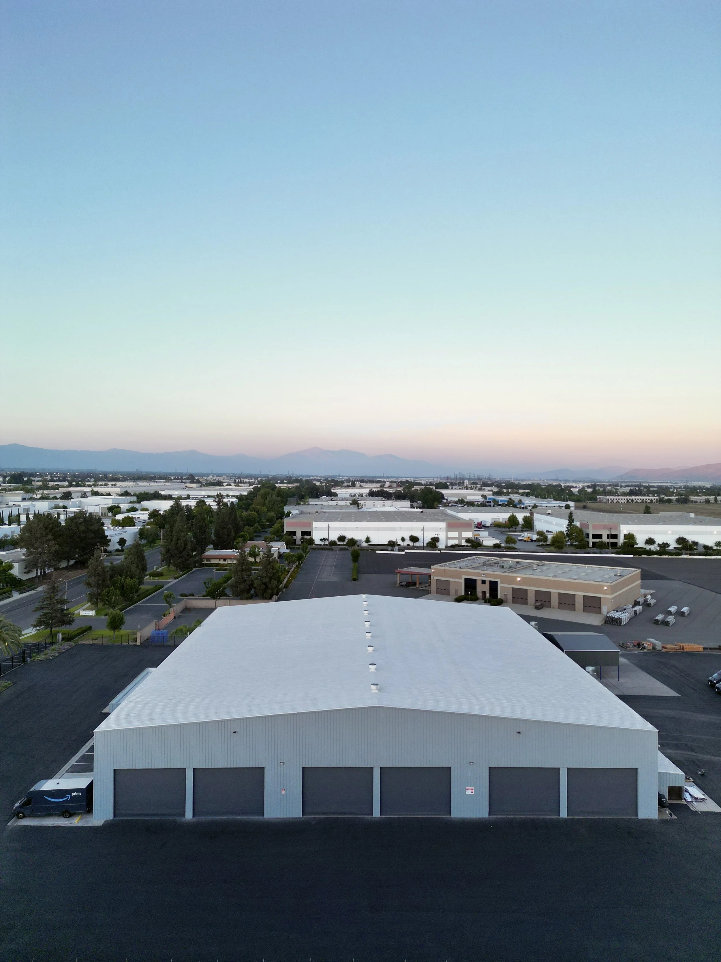 An aerial view of a large metal warehouse building with a large fenced yard for parking and some trees, under a clear sky during sunset in Rancho Cucamonga.