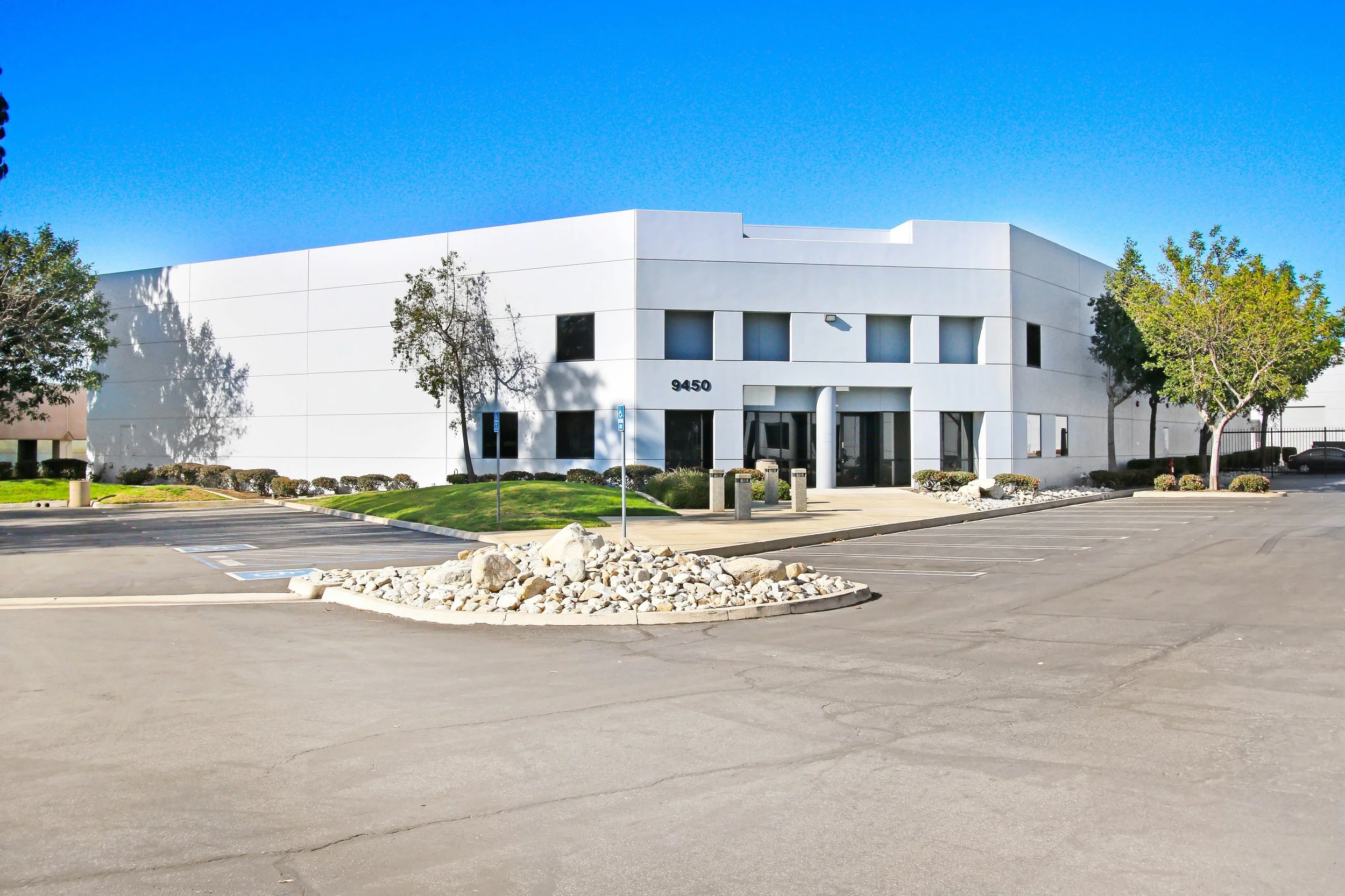 A modern white office building with the number 9450 on the front, surrounded by a parking lot, trees, and small bushes, under a clear blue sky.