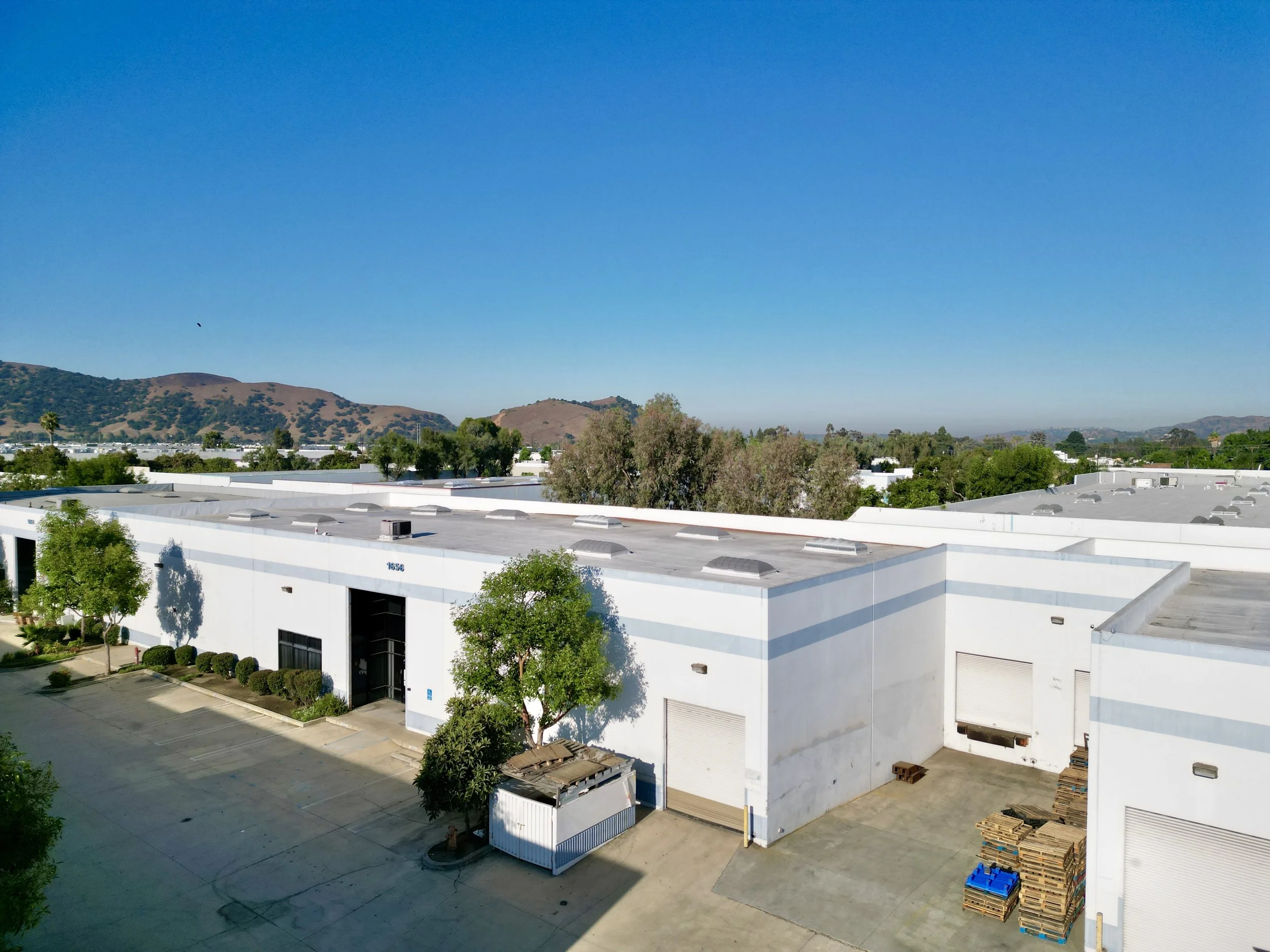 White industrial warehouse building with trees and loading dock area, surrounded by parking lot, under a clear blue sky with distant hills in the background in Pomona.