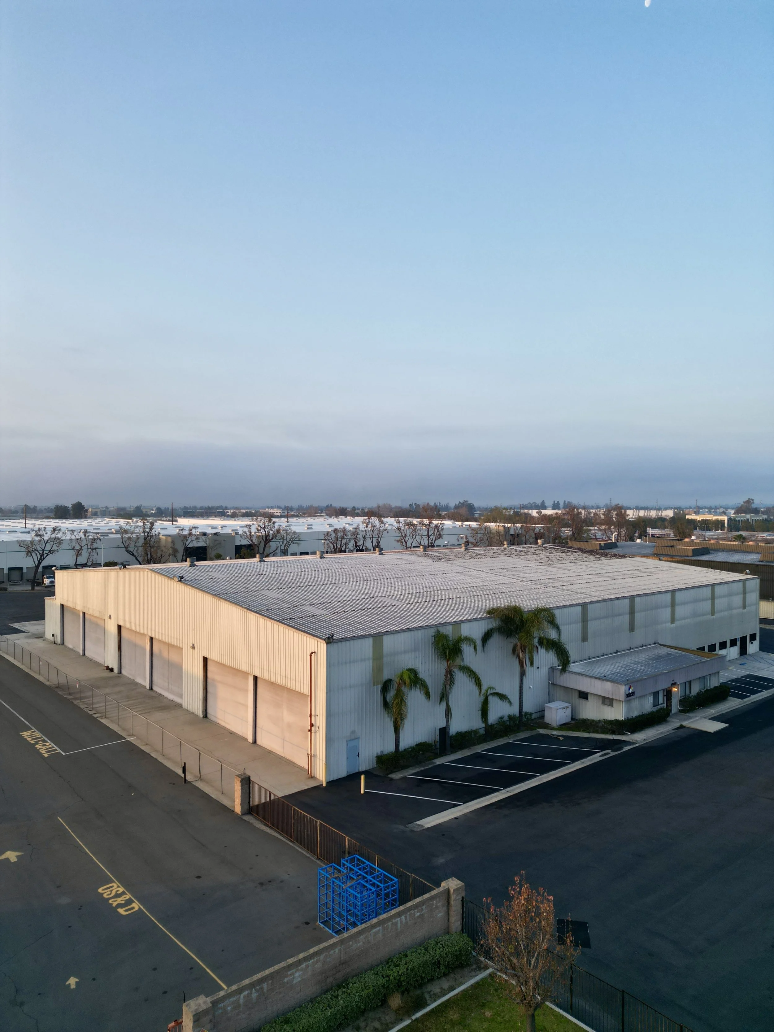 A metal industrial warehouse building with sloped roof, surrounded by parking lot and a few palm trees in front.