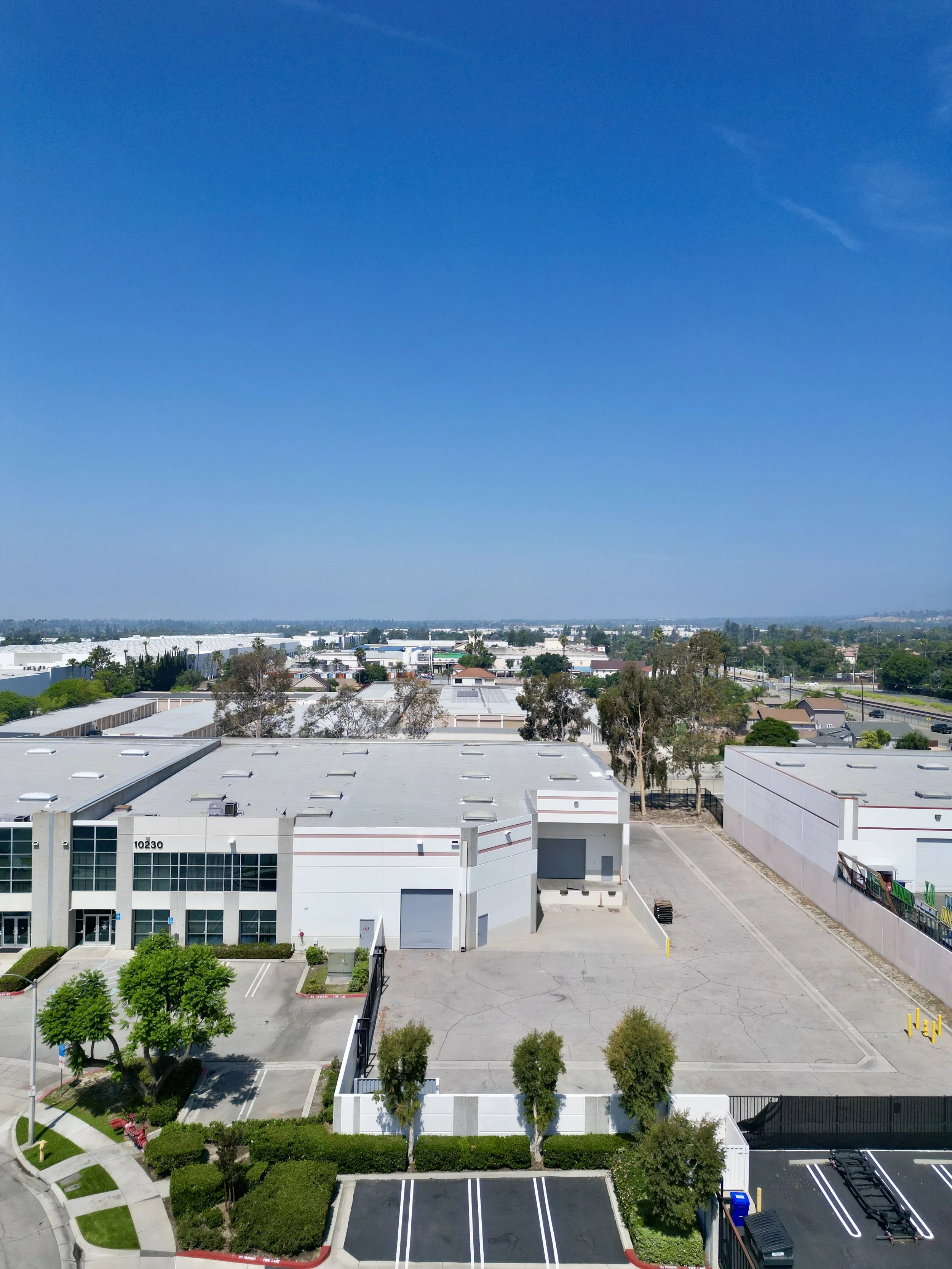 An aerial view of a industrial warehouse building with a private yard, and some trees, under a bright blue sky.