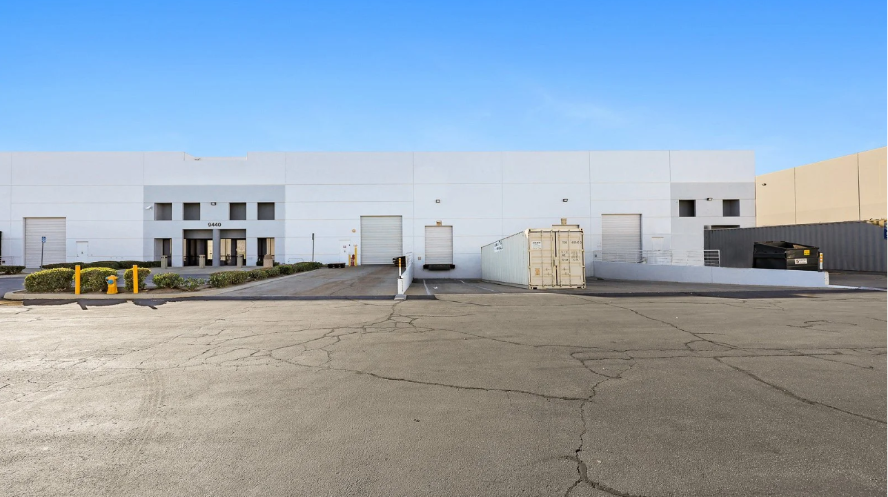 A white industrial warehouse building with three loading docks, a large shipping container, and a dumpster in a parking lot under a clear blue sky.