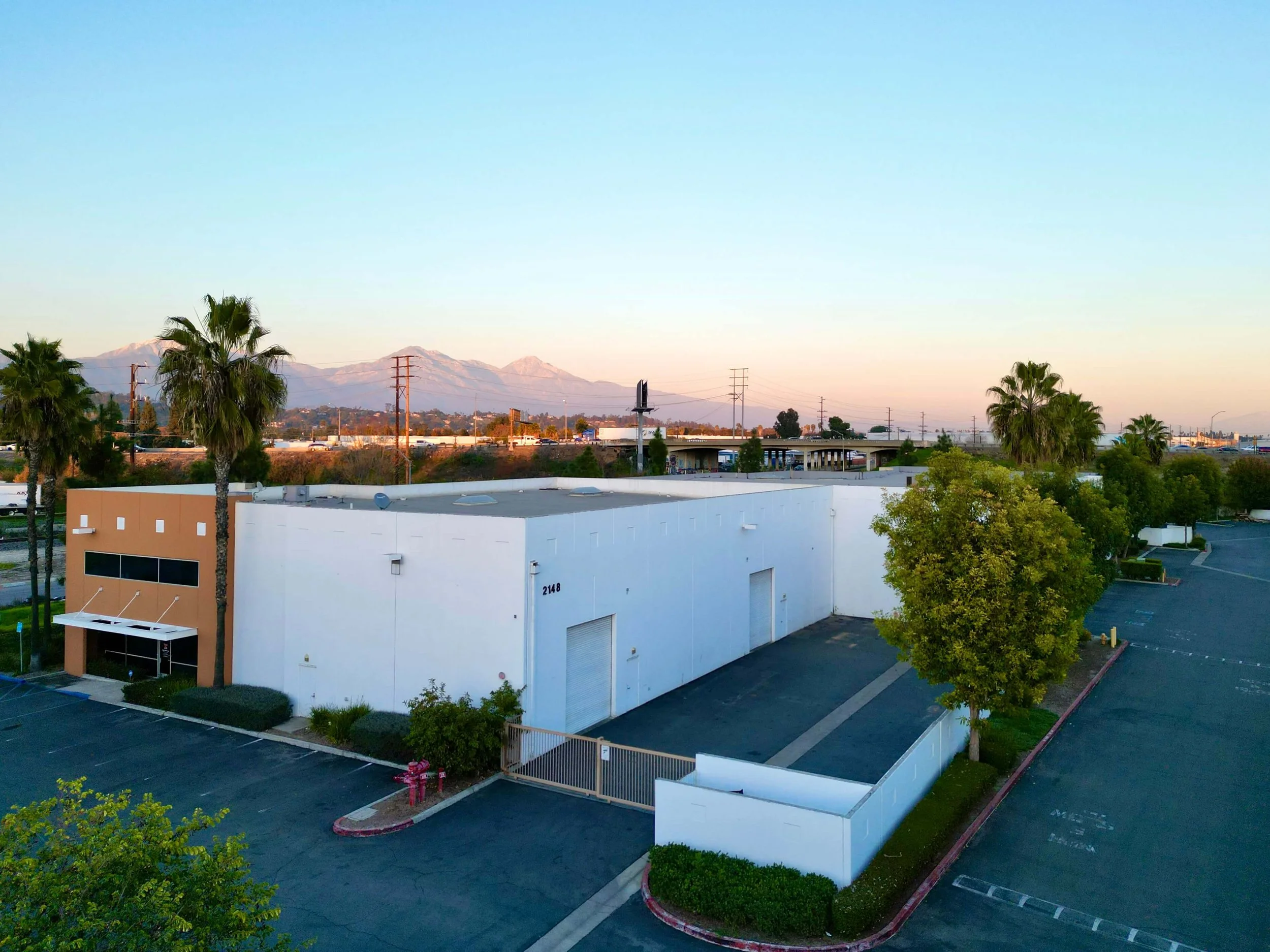 Aerial view of an empty parking lot and a white industrial warehouse with palm trees, green bushes, and a mountain range in the background at sunset.