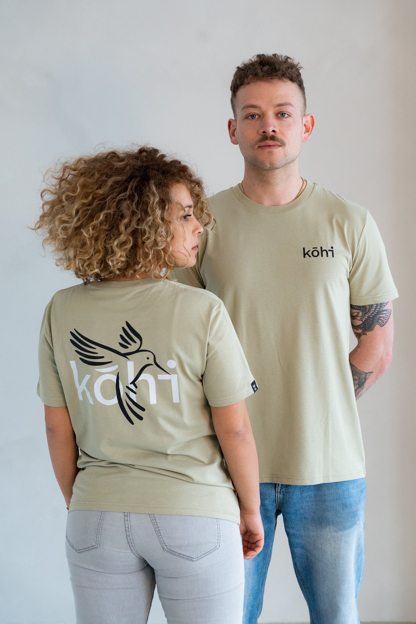 Paar trägt salbeifarbenes Tshirt mit kohi-Logo auf der Brust und kohibri-Druck auf dem Rücken.