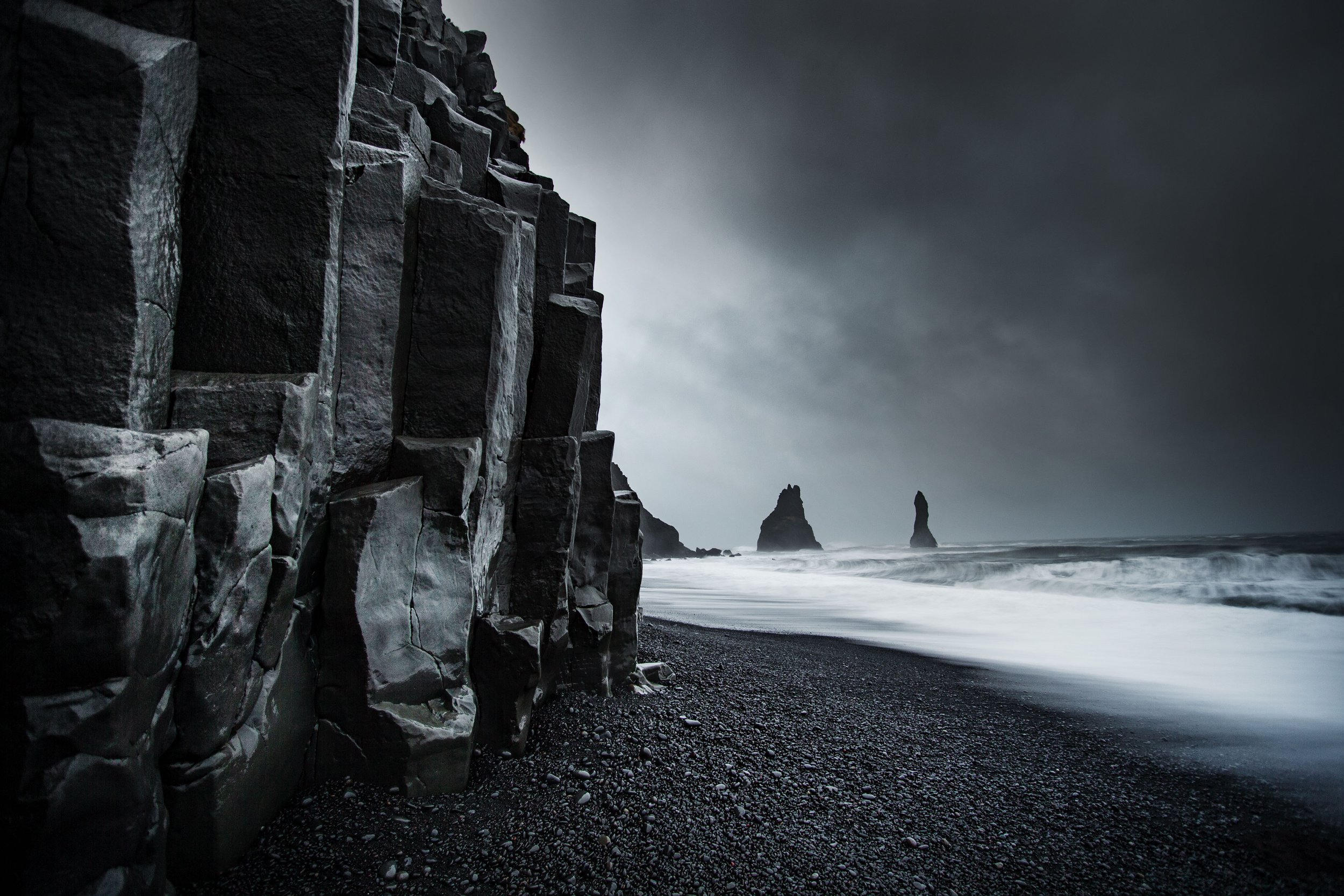 Dark volcanic beach with towering black basalt cliffs, rough ocean waves, and distant sea stacks under cloudy sky.
