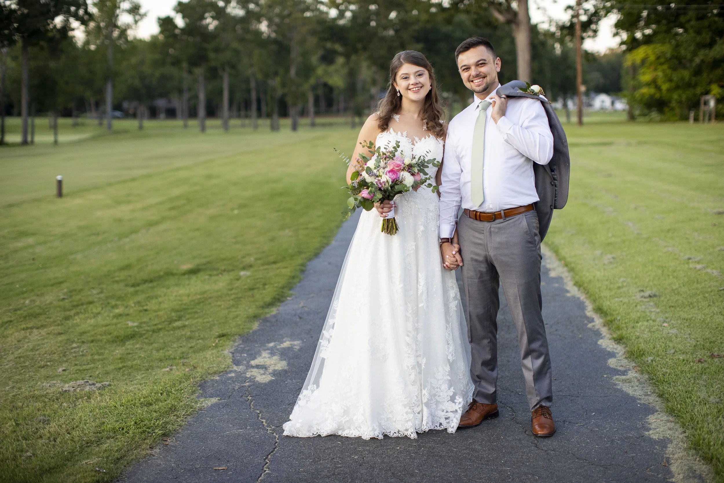 Bride and groom holding hands on a paved path in a park, smiling, with the bride in a white wedding dress holding a bouquet, and the groom in a white shirt, light green tie, and gray pants carrying a suit jacket over his shoulder.