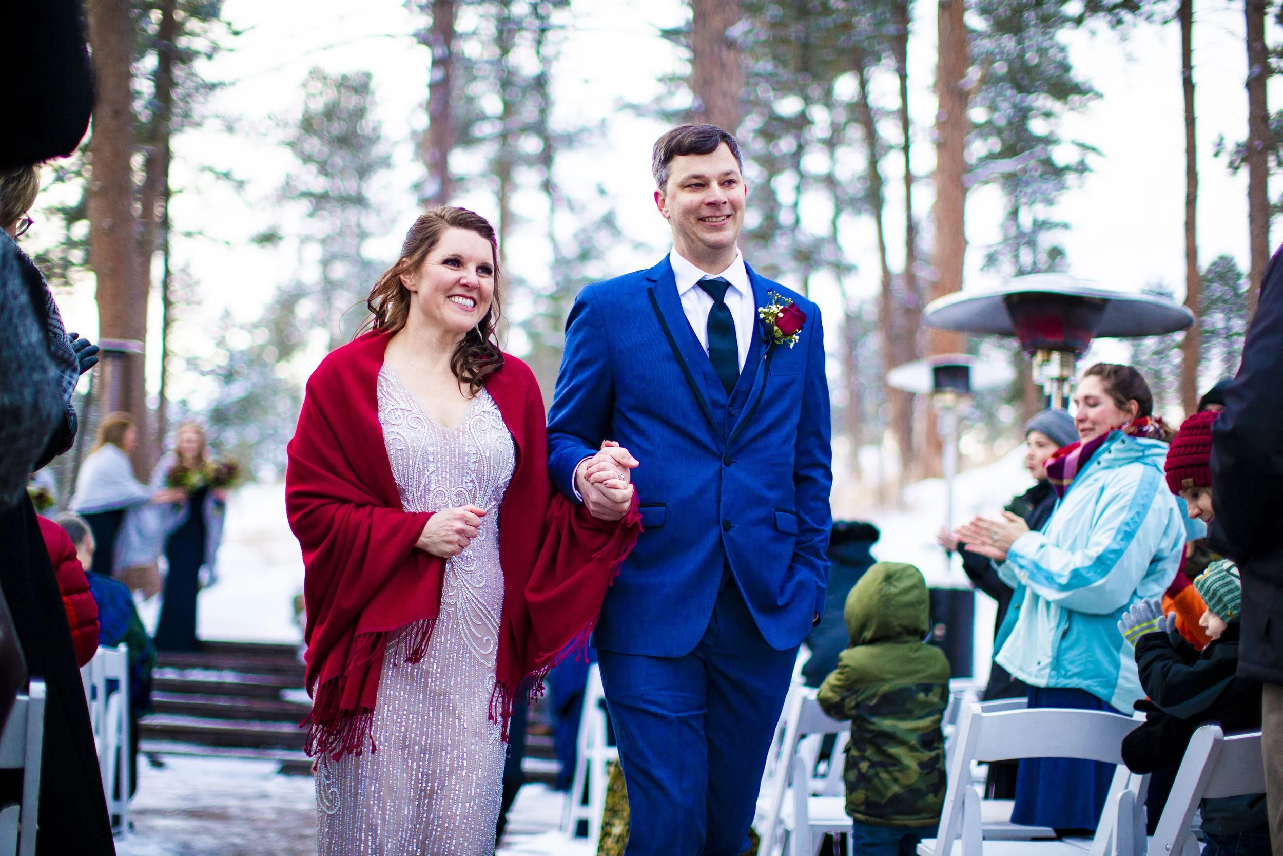 A couple walking down an aisle outdoors in winter, holding hands, during their wedding ceremony, with guests clapping and celebrating around them.