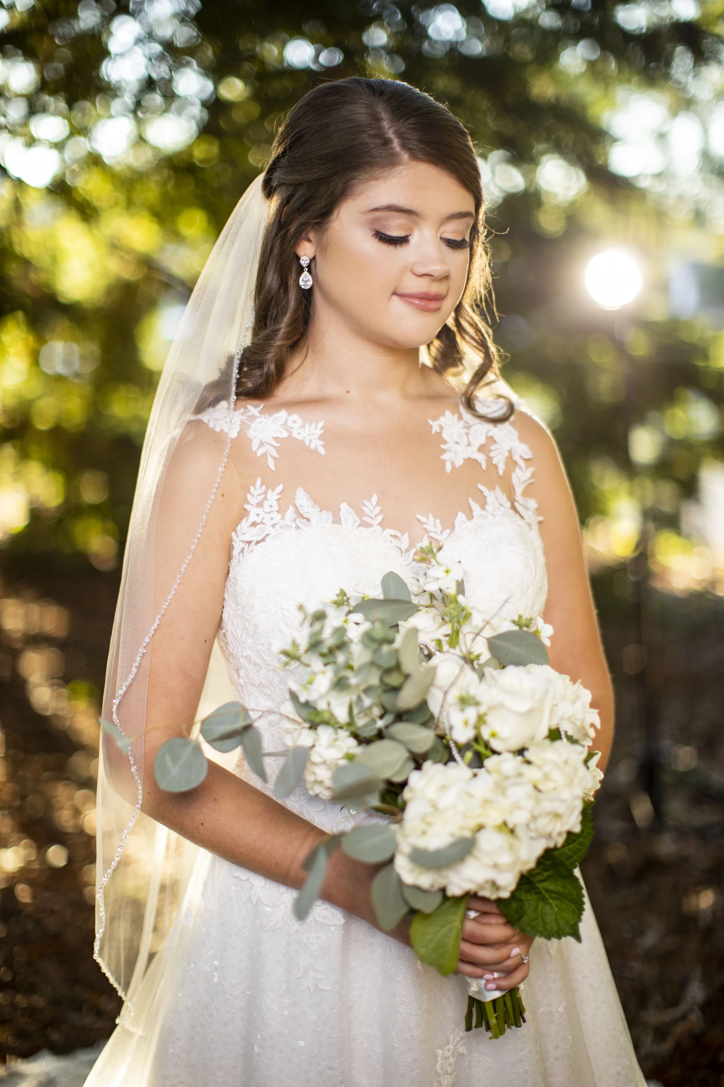 Bride in a lace wedding dress holding a bouquet of white flowers and greenery, standing outdoors with sunlight filtering through trees in the background.