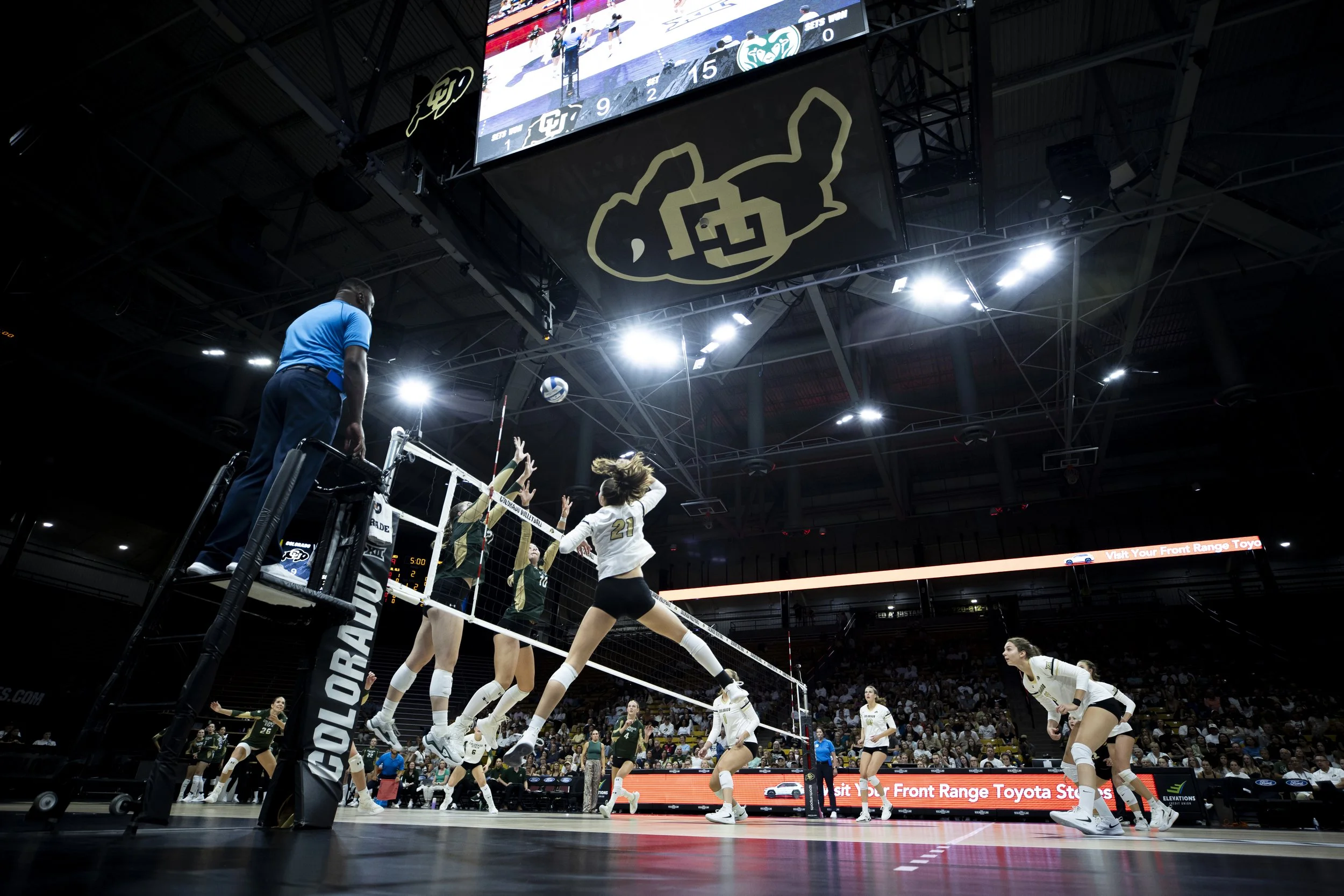 Indoor volleyball game with players jumping to hit the ball over the net, referee standing on a platform, and spectators in the background.