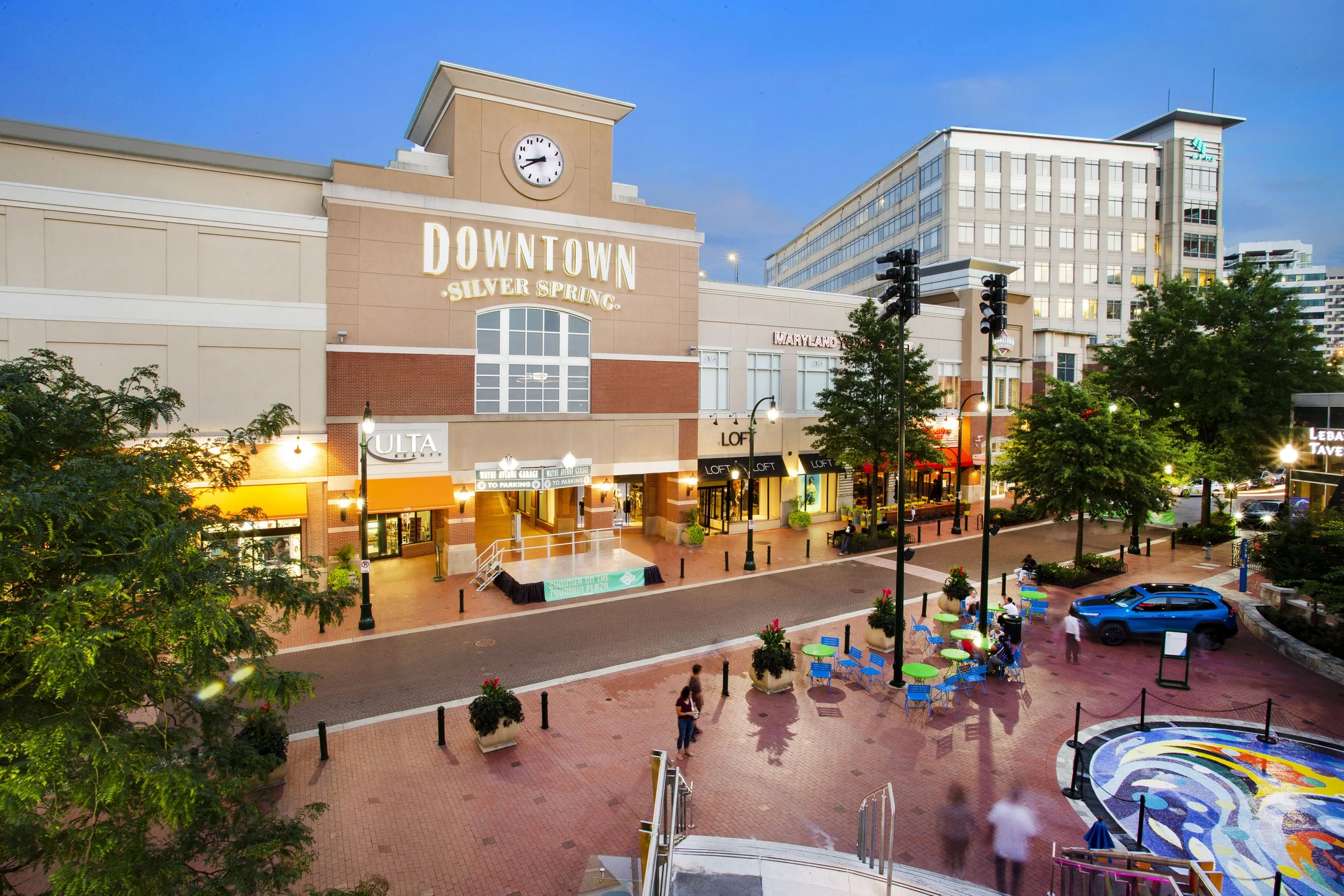 City street view at dusk with shopping center, trees, outdoor seating, parked car, and people walking