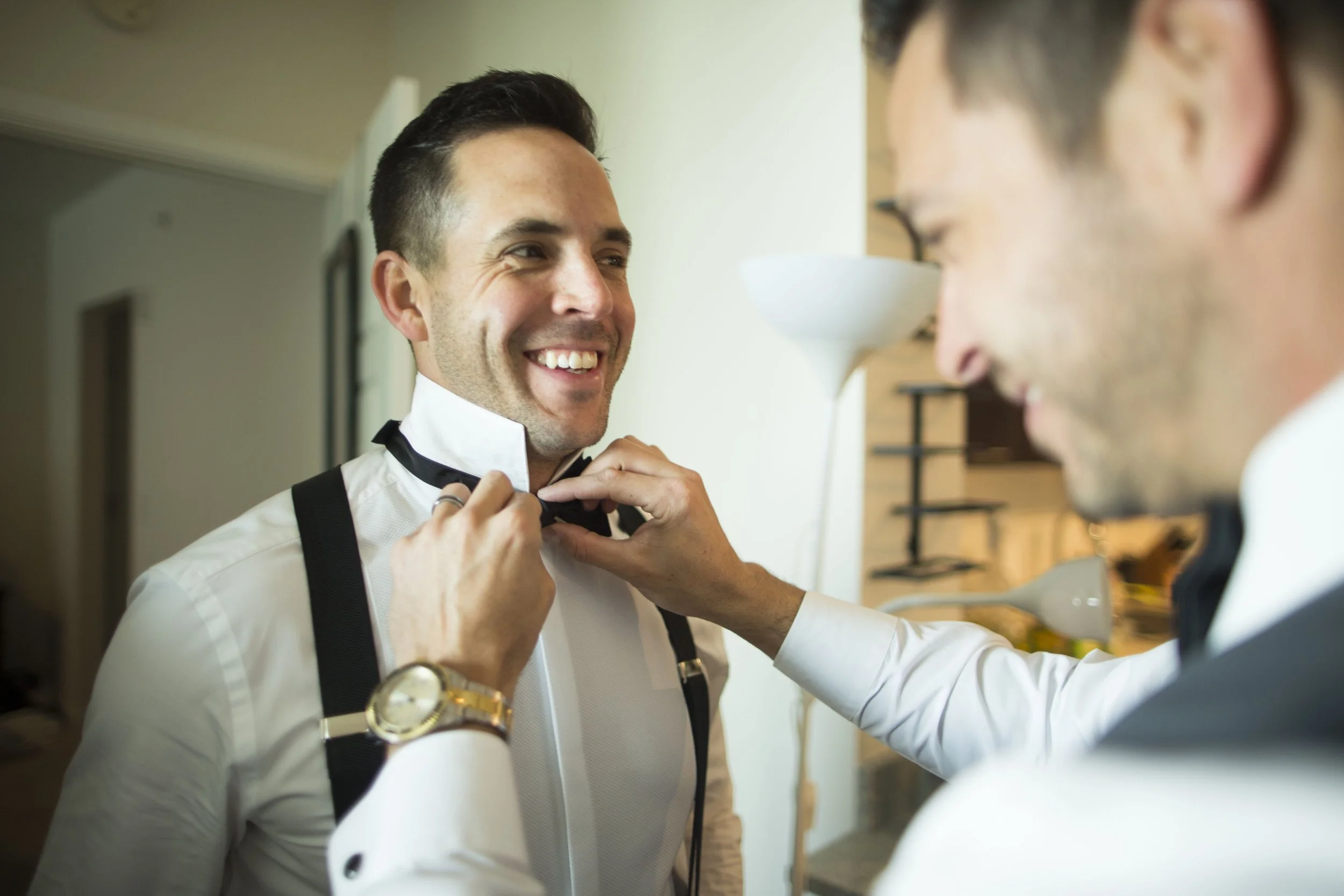 Two men in formal attire, one helping the other adjust a black bowtie, smiling warmly in a warmly lit room.