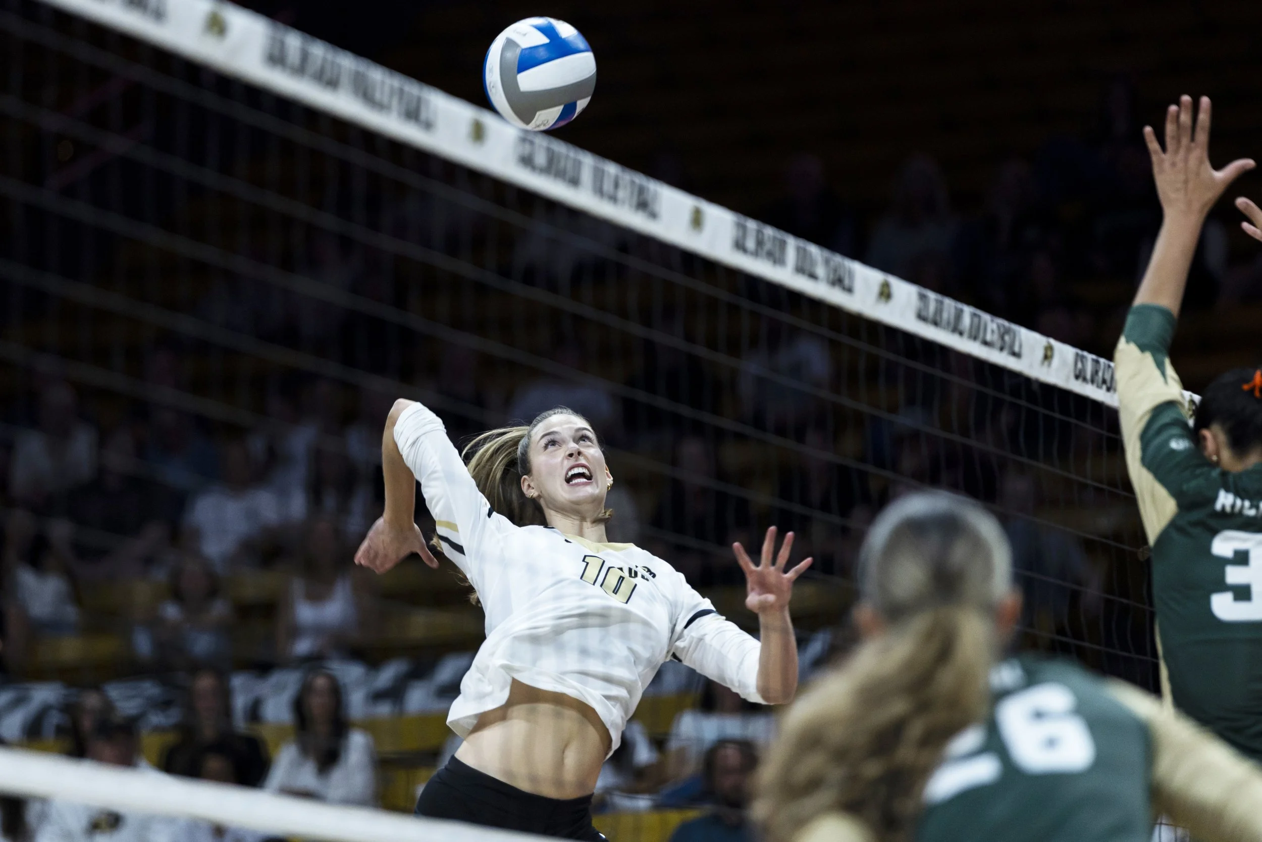 A volleyball player in a white jersey is jumping to hit the ball over the net, with her face showing effort. Opponent players in green jerseys are preparing to block.