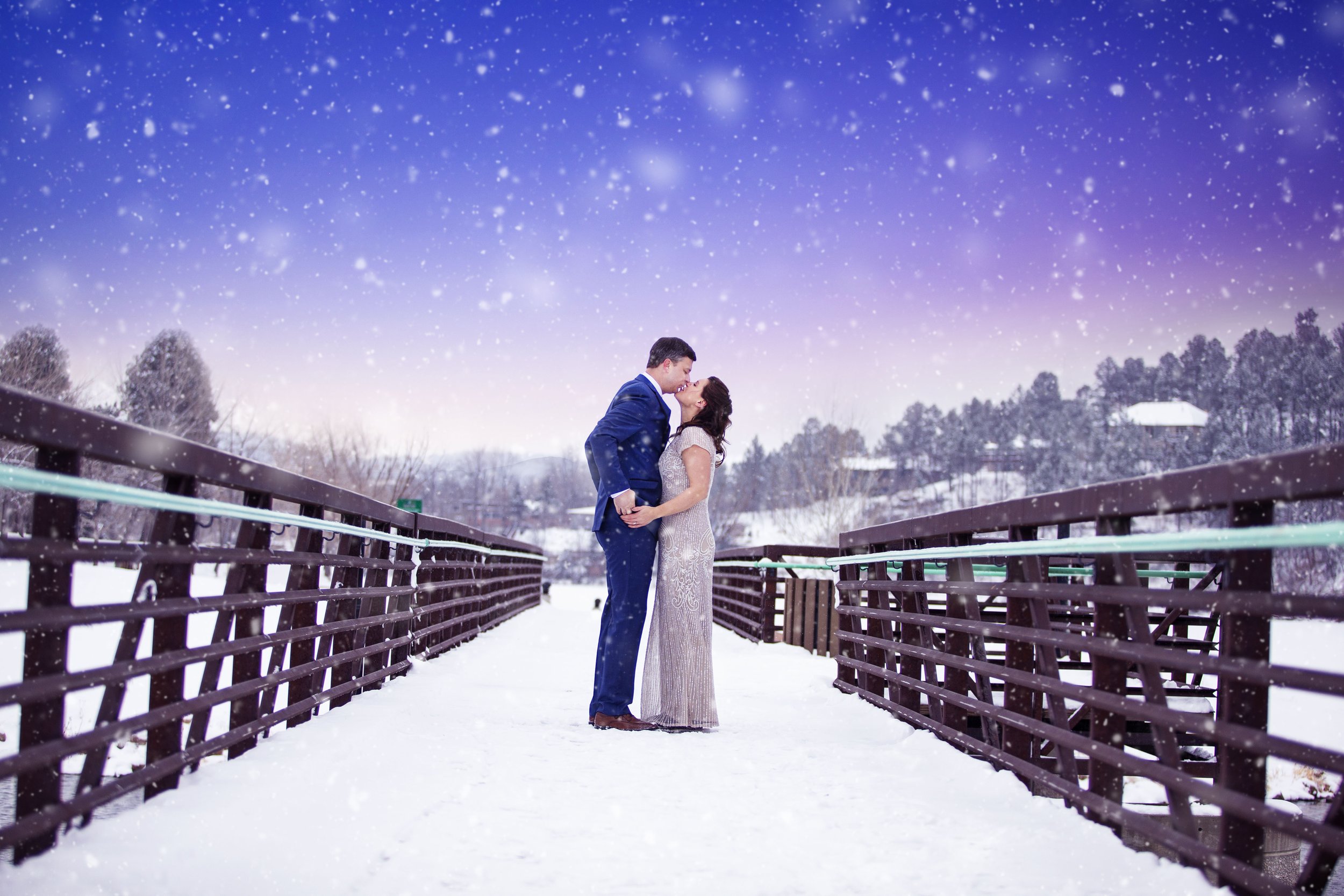 A couple in formal attire sharing a kiss on a snowy bridge with falling snow and a winter landscape in the background.