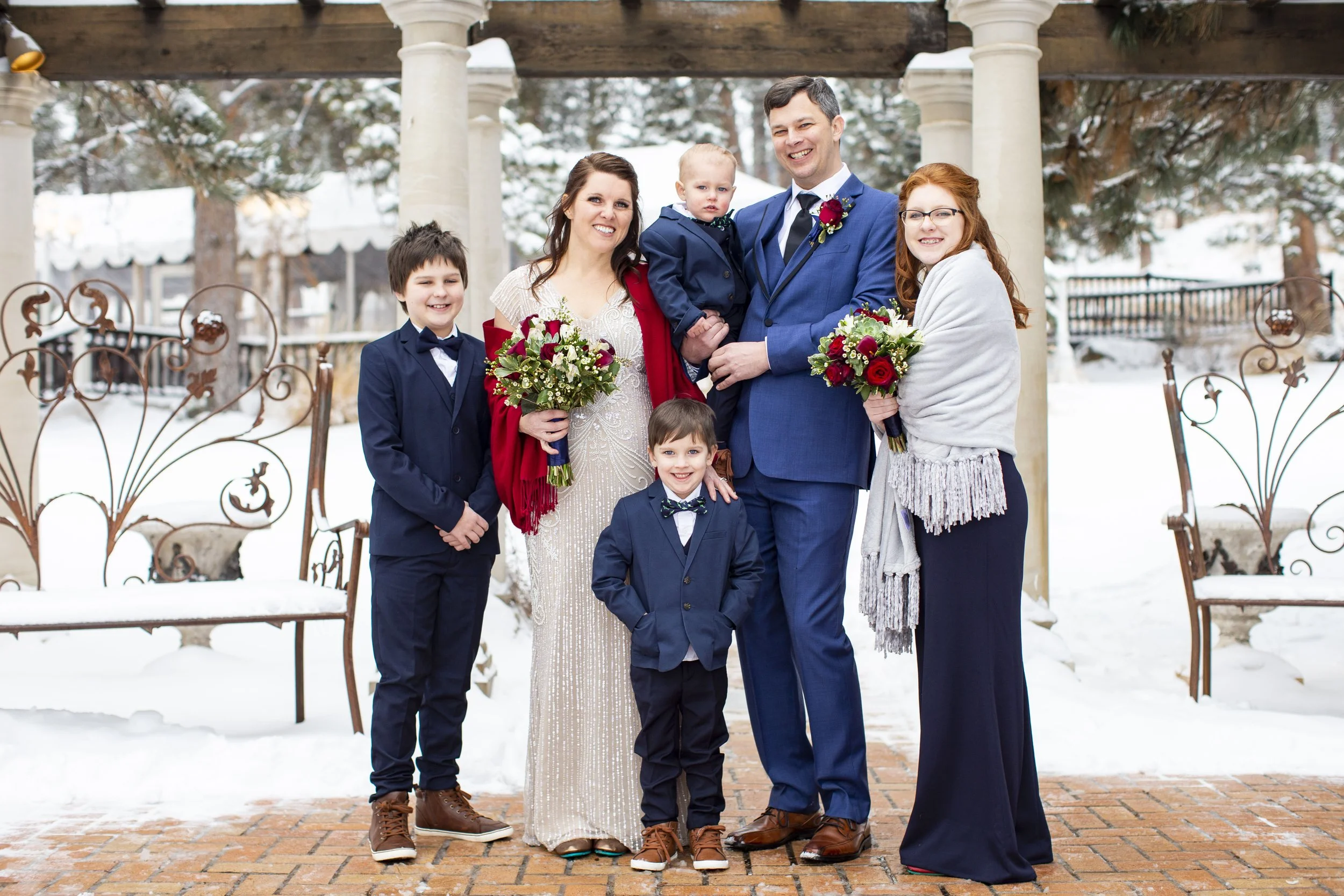 A wedding party standing outdoors in snowy weather, with three women, two men, and three children, all dressed formally, some holding bouquets of red and white flowers, smiling in front of a garden gazebo with snow-covered trees in the background.