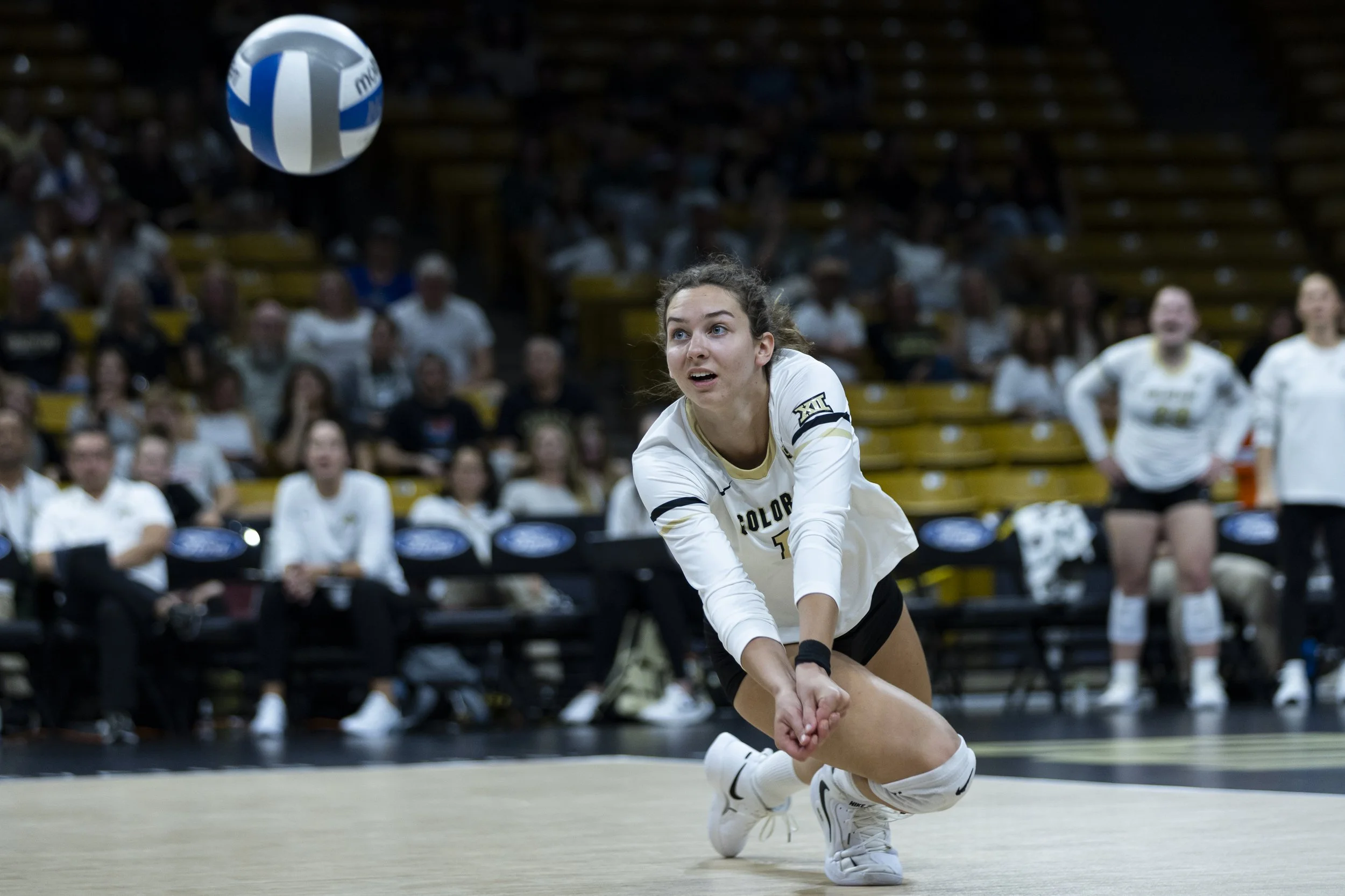 A female volleyball player is diving to her right to dig the ball during a match, with spectators seated in the background.