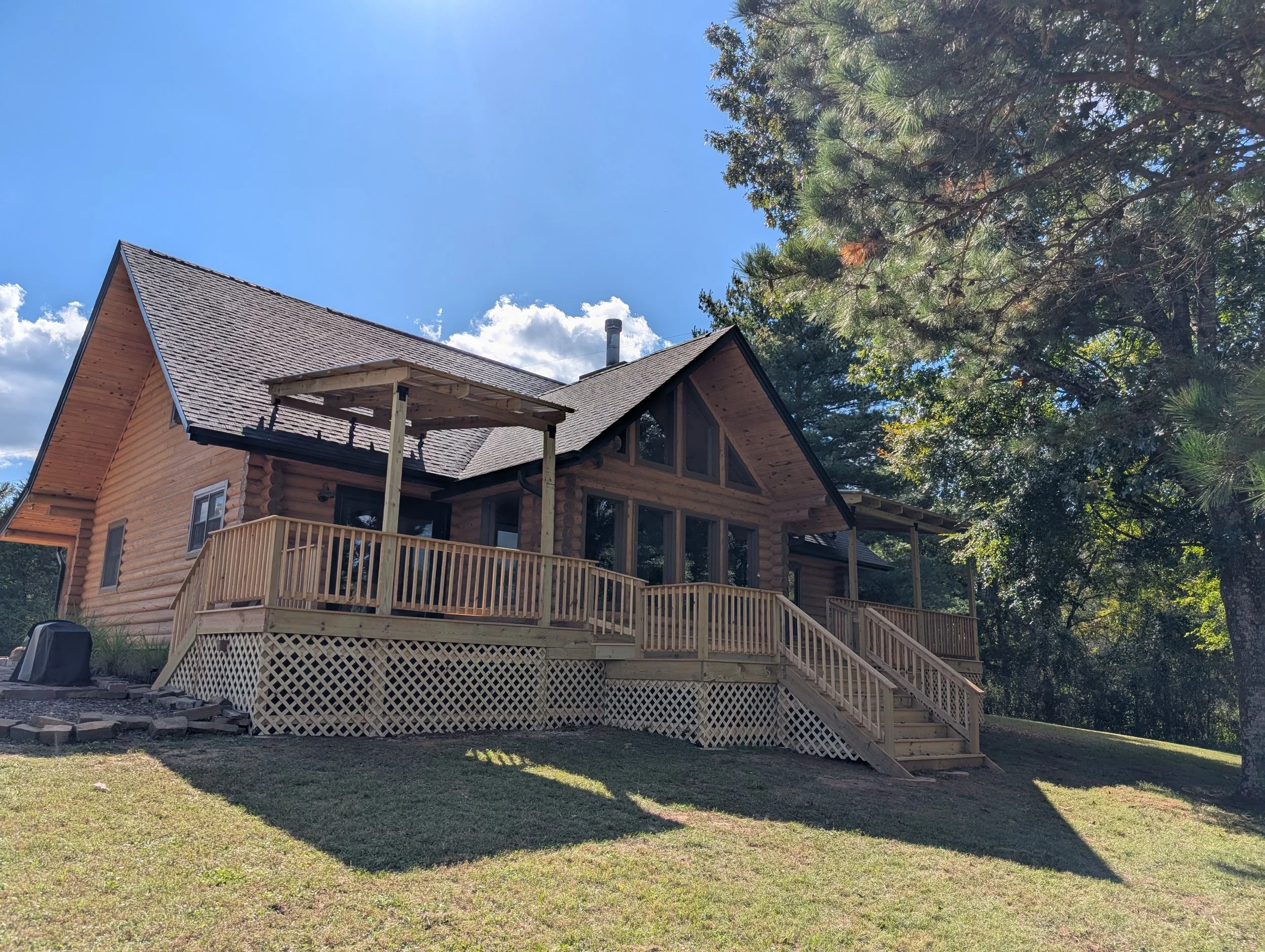 A new wooden house with a spacious wraparound deck, surrounded by grass and trees, under a blue sky with clouds.