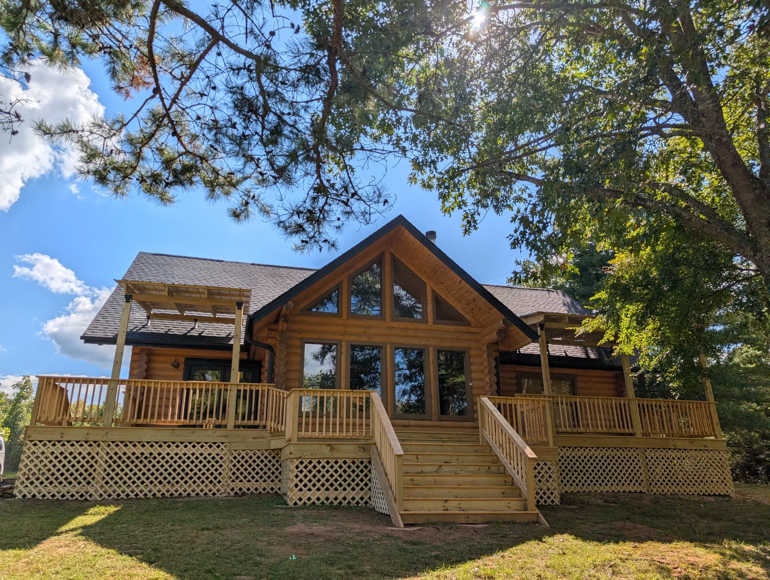 A wooden house with large glass windows, a deck with railings, and steps leading up to it, surrounded by trees and under a blue sky with clouds.