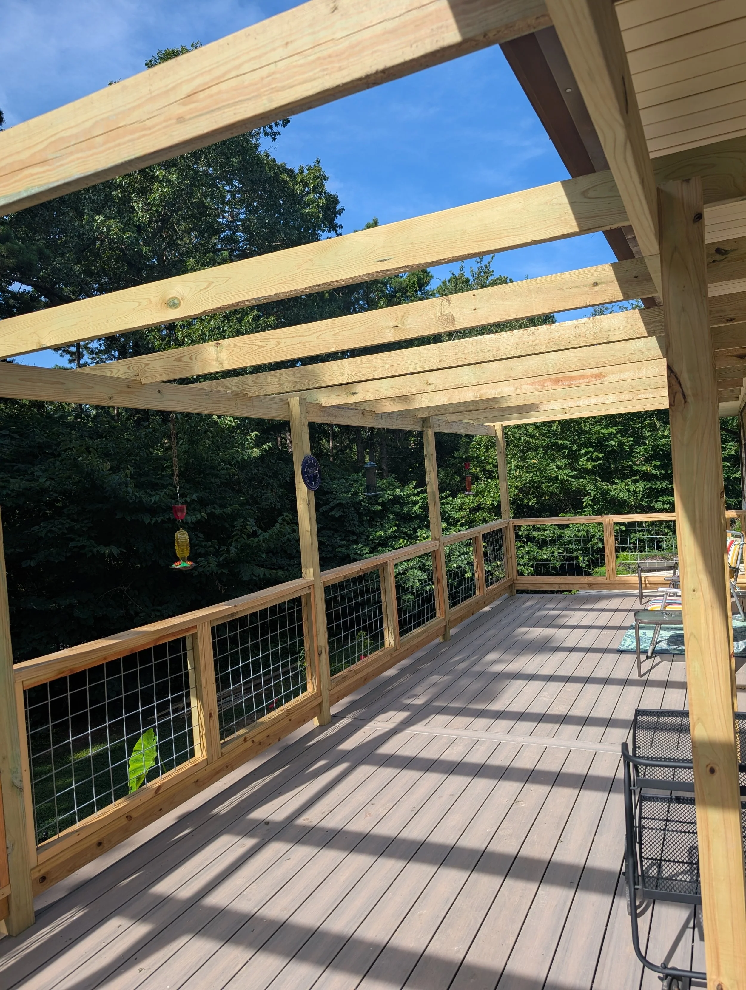 Wooden deck under construction with partial roof frame, surrounded by a forested area, and outdoor furniture on the deck.