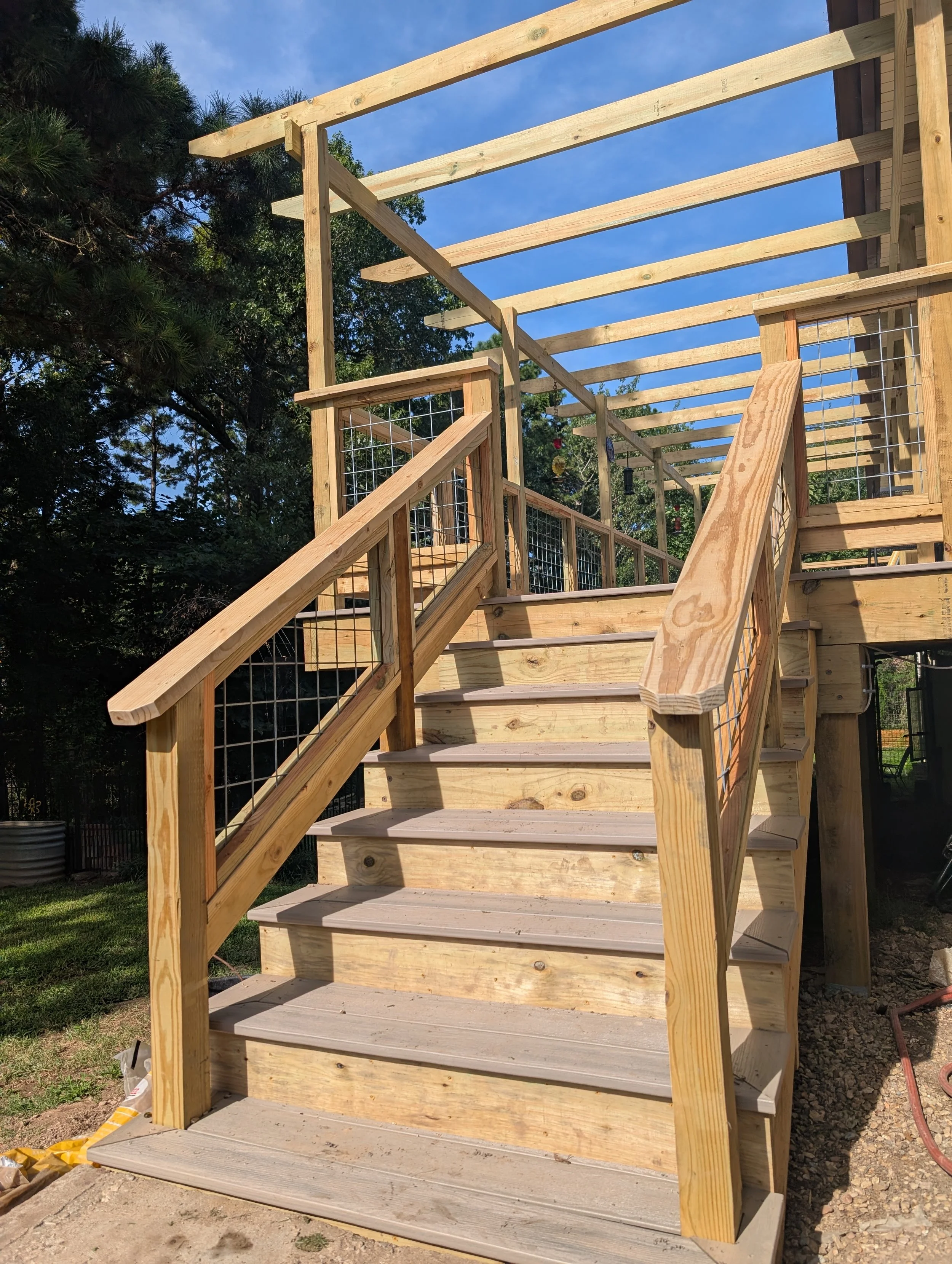 Wooden staircase with handrails under construction, leading up to an outdoor deck, with a partially built roof frame and trees in the background.