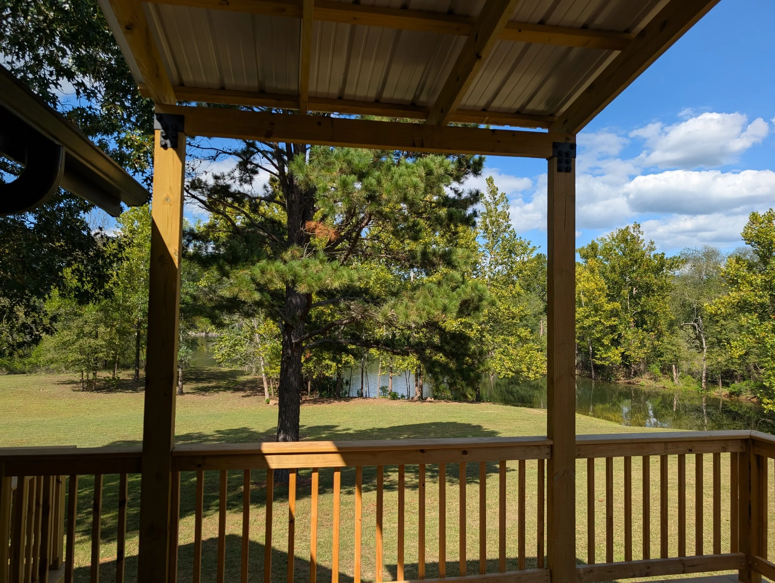 View from a wooden deck looking at a grassy yard, trees, and a small body of water under a partly cloudy sky.