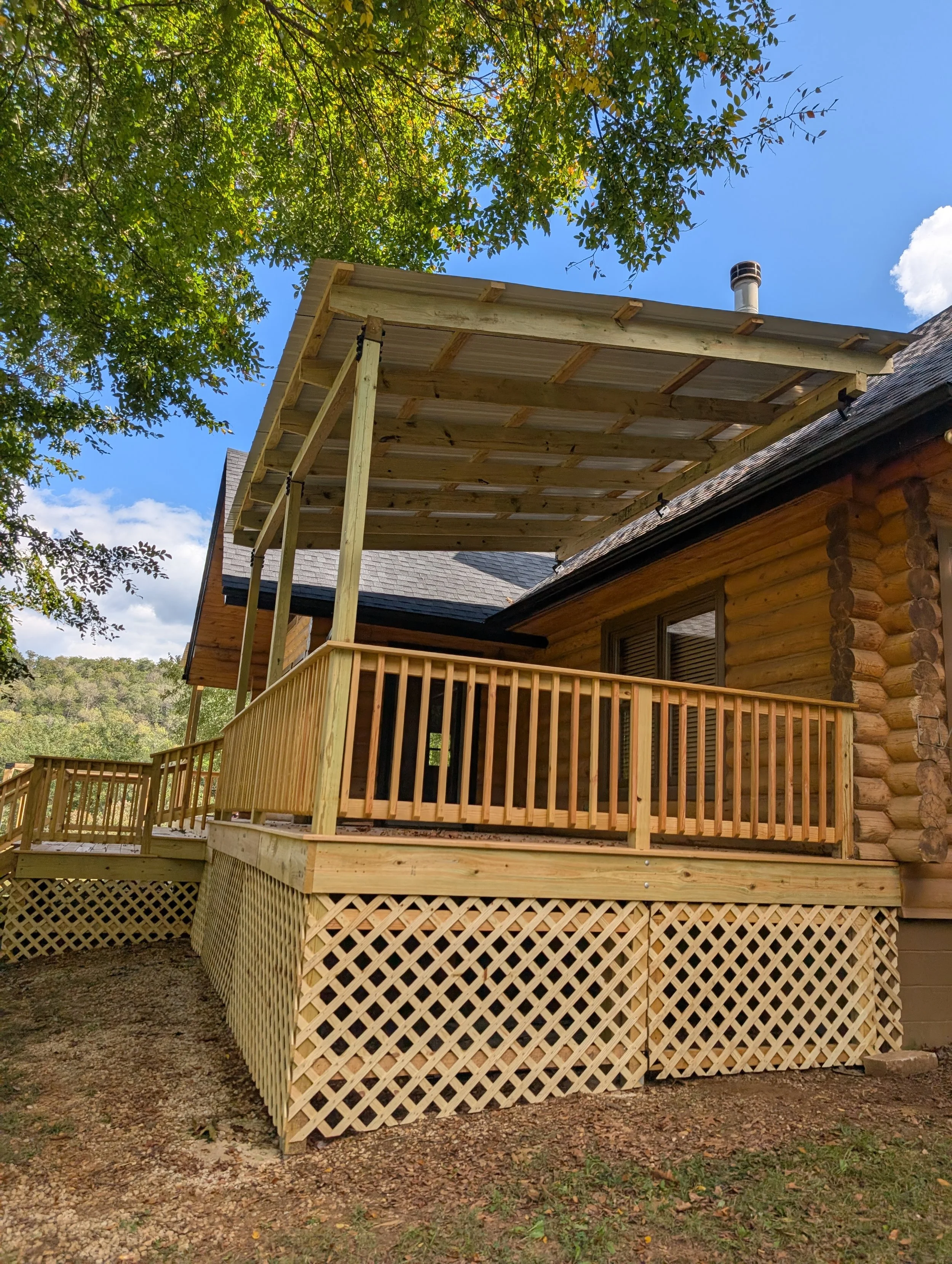 Wooden deck with railing attached to a log cabin, with an overhead roof extension under construction, surrounded by trees and a blue sky.