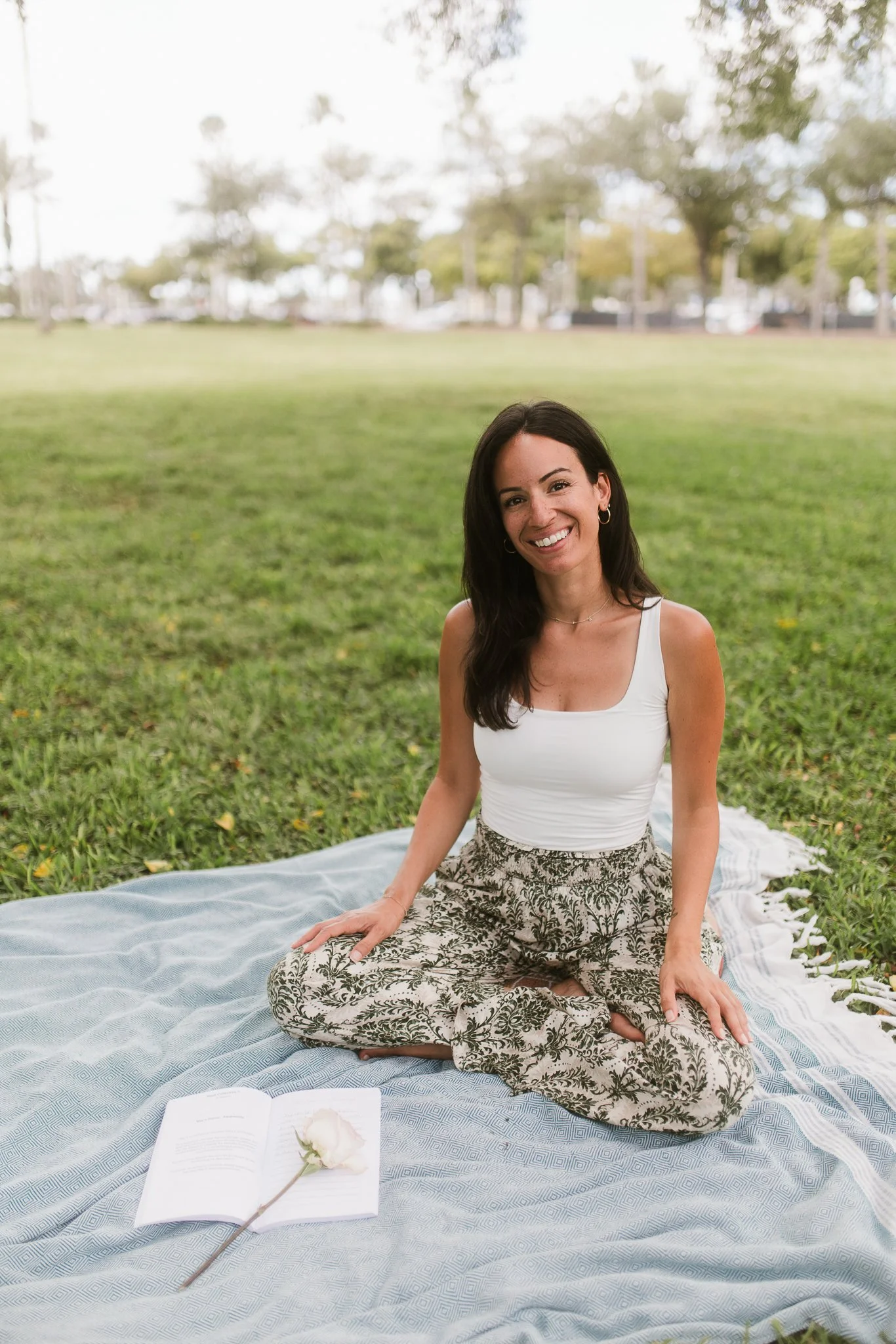 A smiling woman sitting cross-legged on a blanket in a park, with an open book and a white flower in front of her. She is wearing a white tank top and patterned pants, with trees in the background.