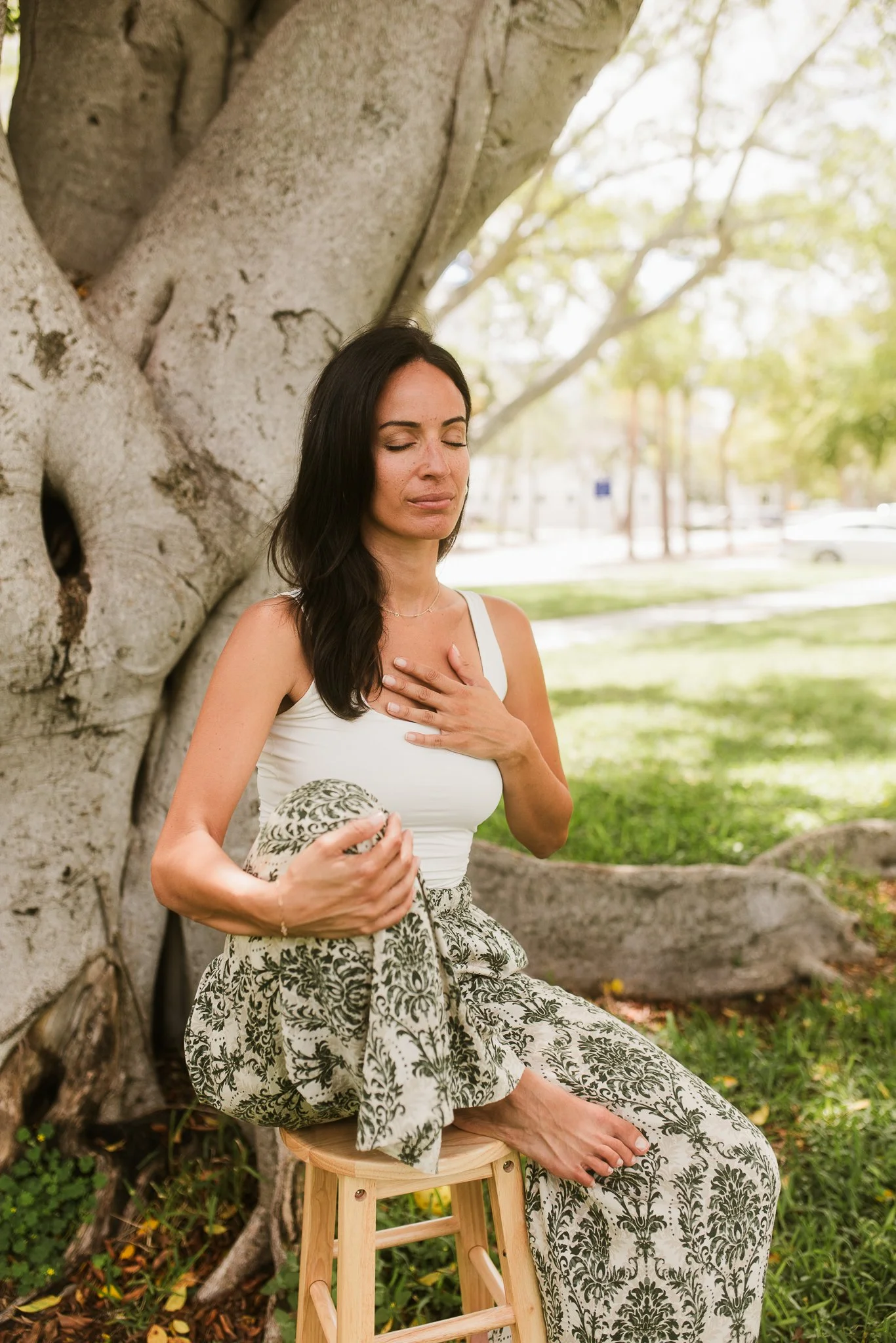 A woman with long dark hair and a white top, sitting outdoors against a large tree, with her eyes closed and one hand on her chest, holding a patterned cloth in her lap, in a peaceful moment surrounded by greenery and sunlight.