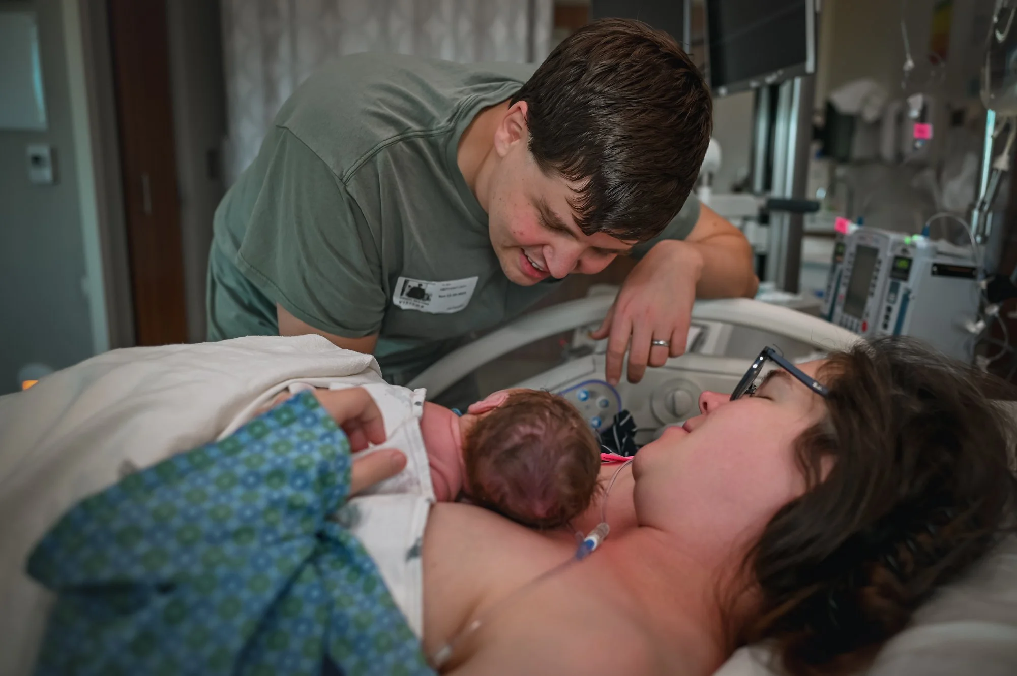 A woman lying in a hospital bed with a newborn baby on her chest, a man leaning over her, all in a hospital room with medical equipment in the background.