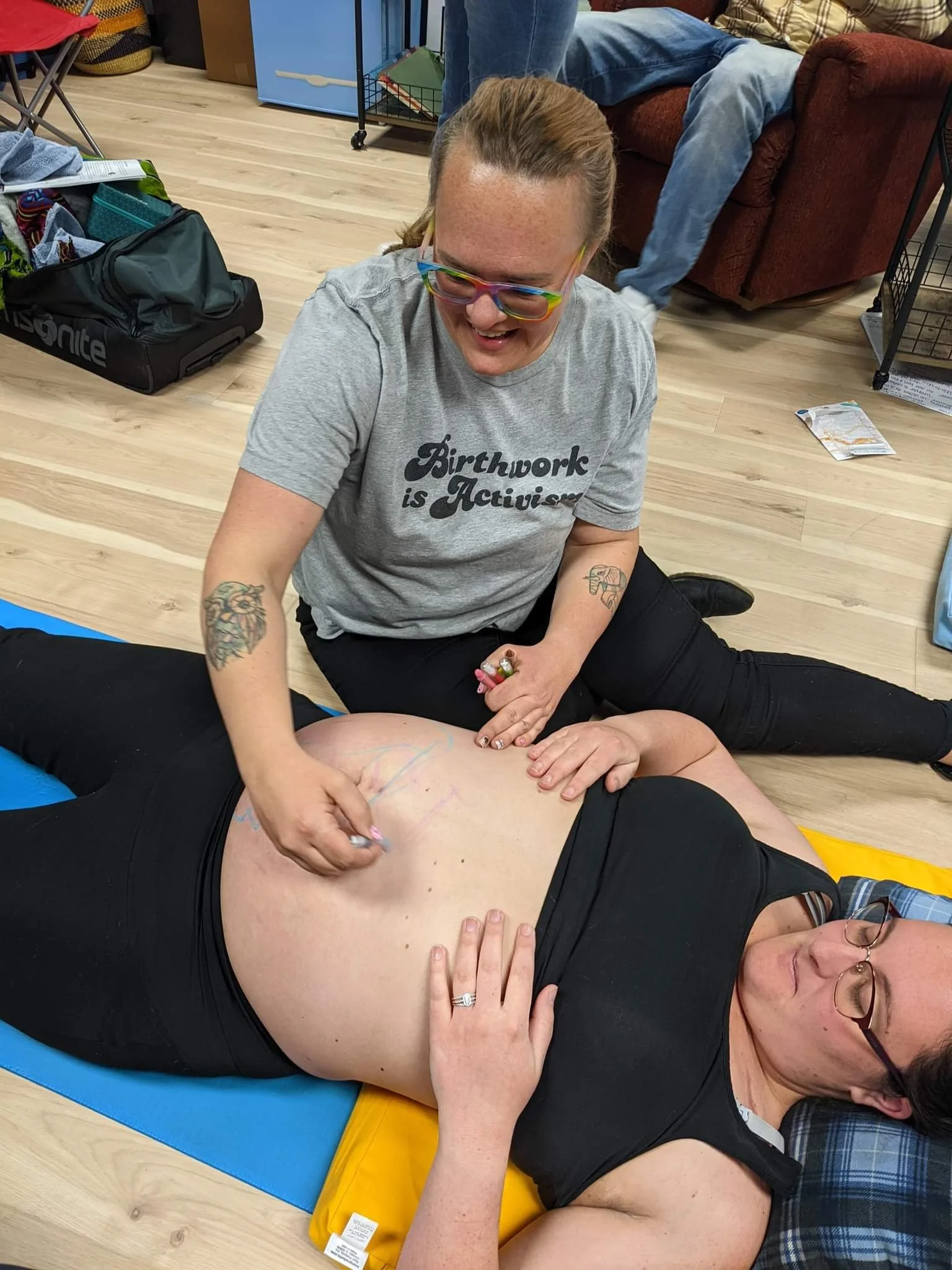 A woman with tattoos and colorful glasses is giving a tattoo to a man lying on the floor. The woman is smiling and wearing a gray t-shirt that reads 'Birthwork is Activism'. The man is lying on a yellow and blue mat with his shirt pulled up, exposing his stomach.