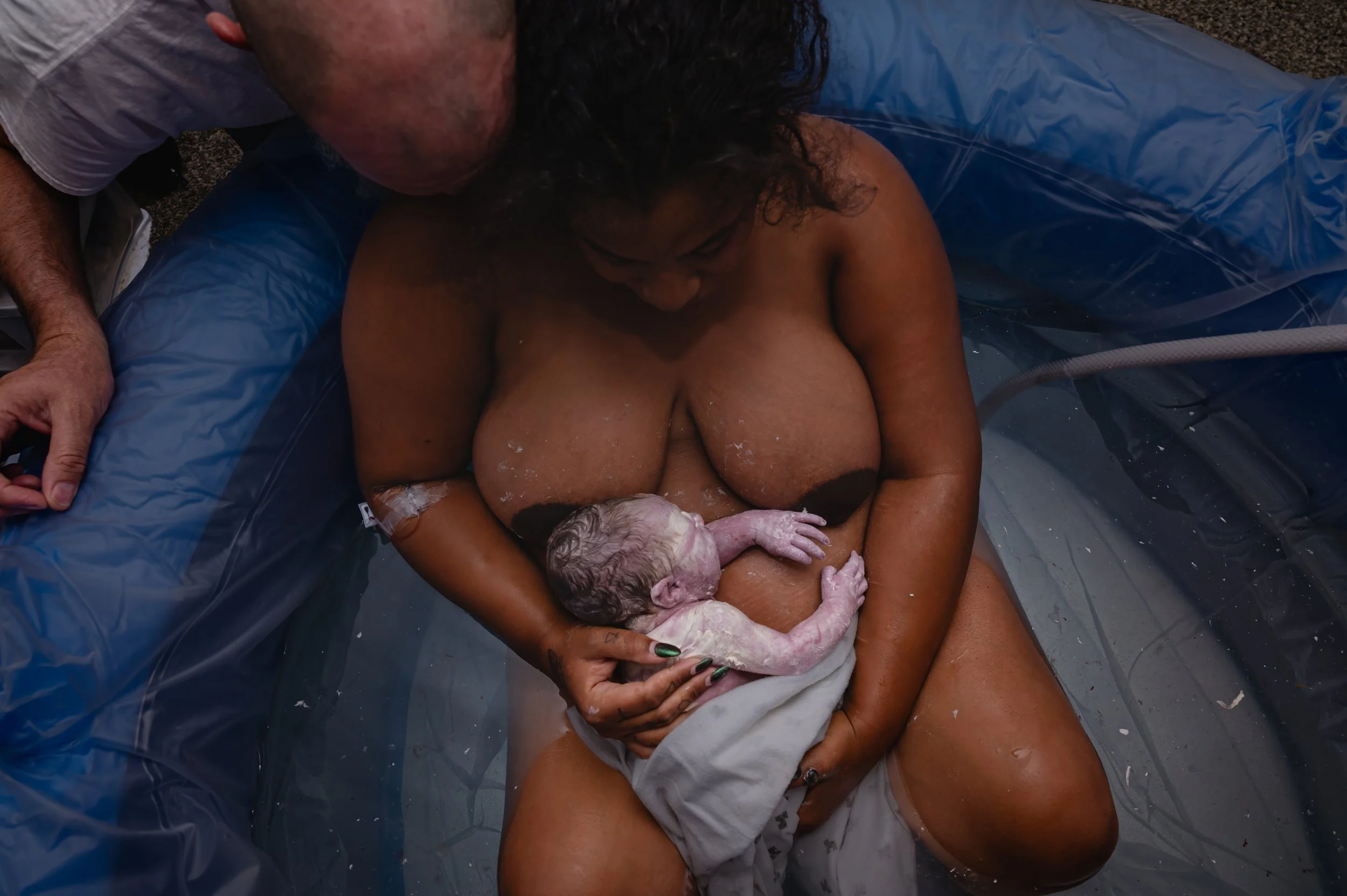 A woman giving birth in a water birth pool, holding a newborn baby with the assistance of a person leaning over her.