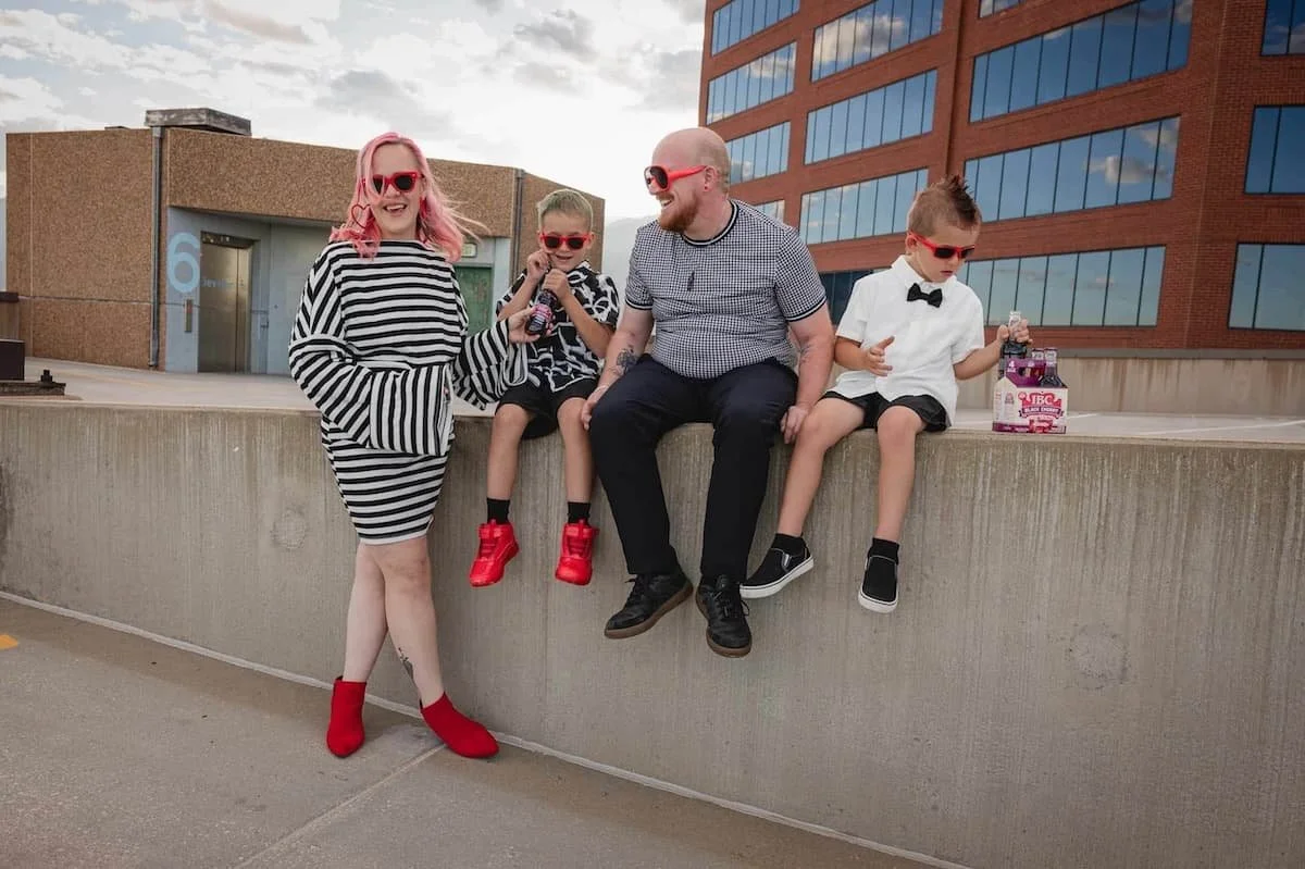 A family of five sitting and standing on a concrete ledge outdoors, with modern buildings in the background. They are all wearing stylish clothing and red sunglasses, and appear to be enjoying themselves.