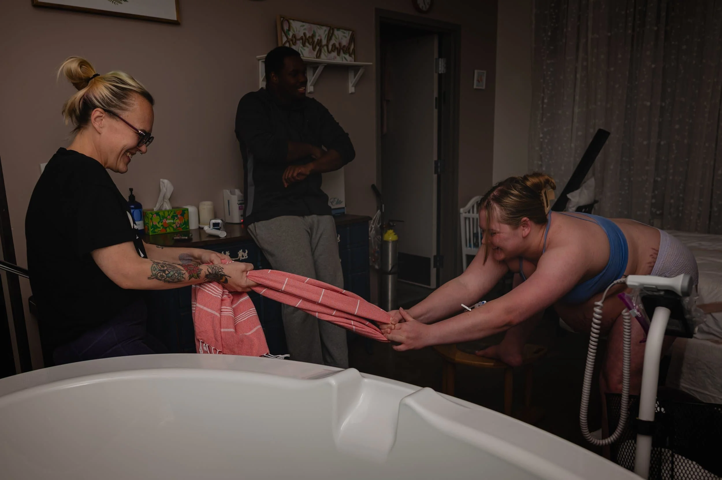 A woman in labor hands a towel to a nurse while a man and another woman watch. The woman is smiling and leaning forward on a hospital bed, preparing for childbirth in a hospital room.