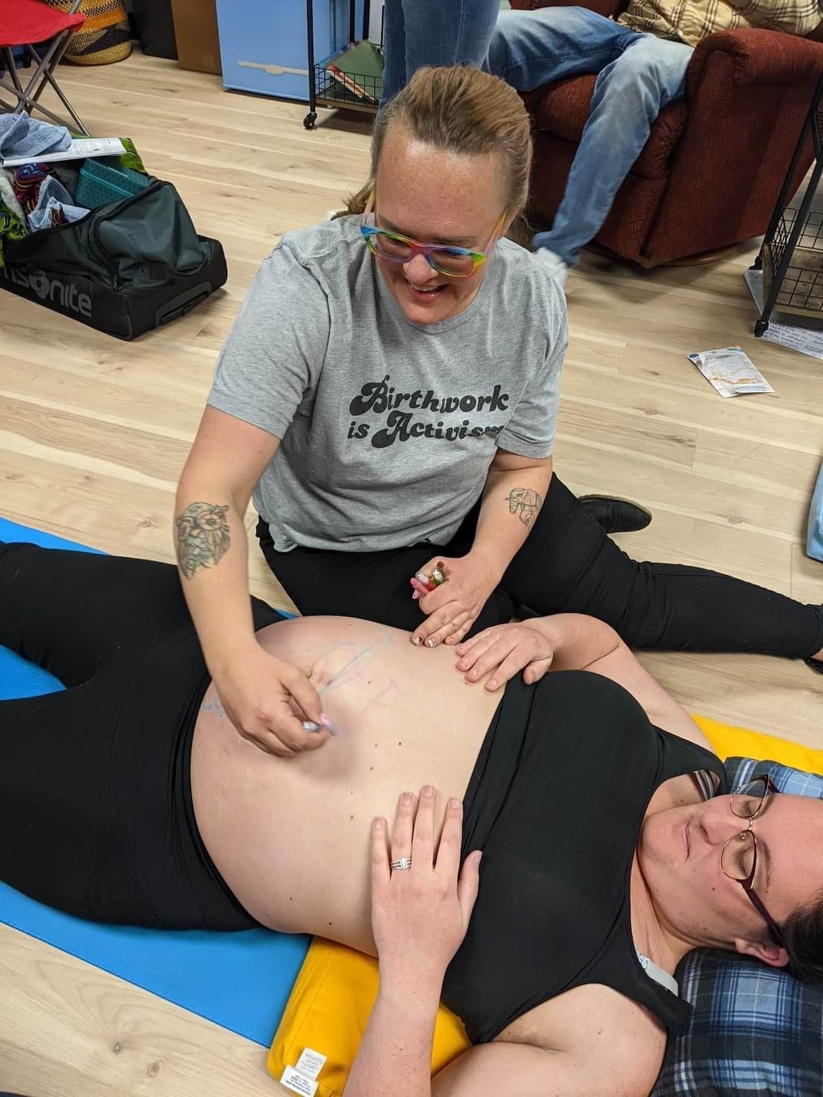 A birth doula smiles while drawing on a pregnant belly, both looking happy and relaxed indoors on a mat.