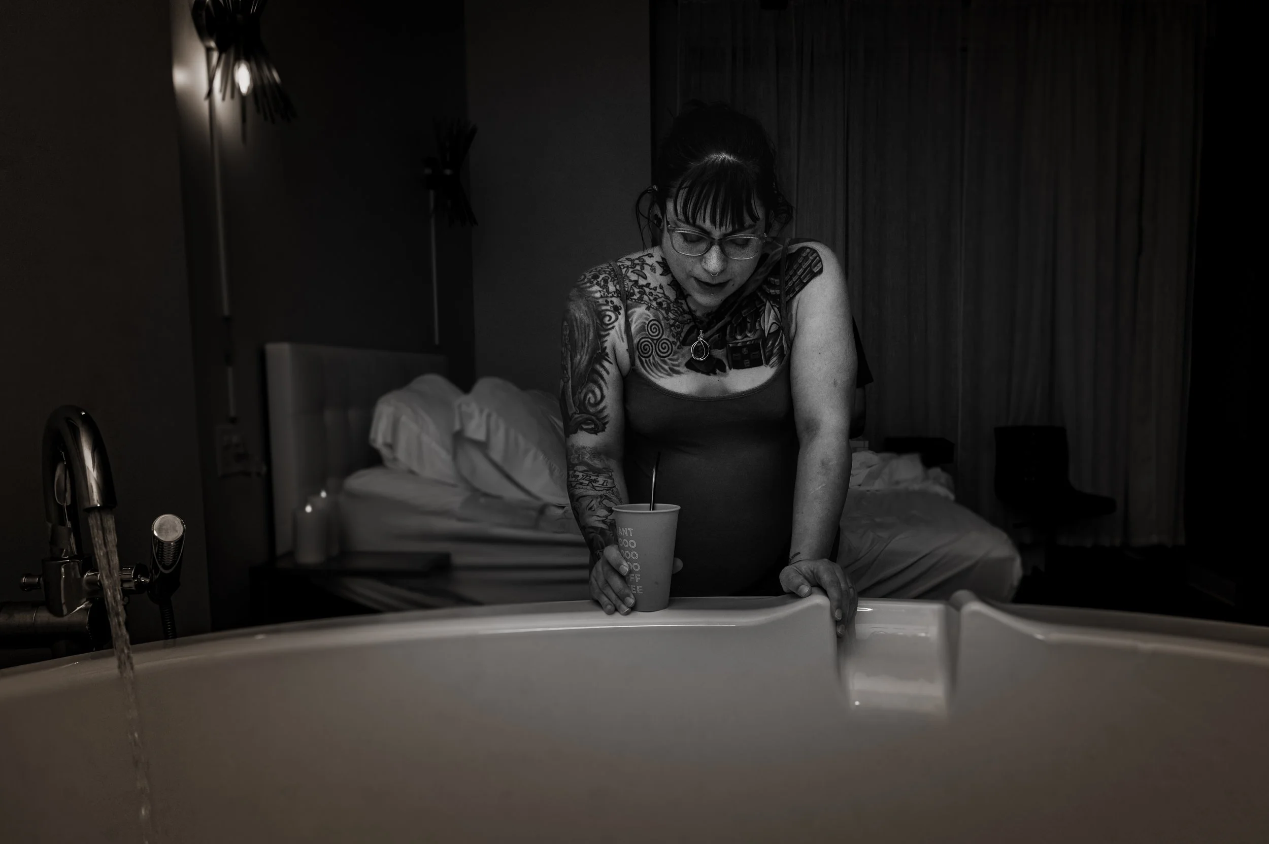 A woman with tattoos and glasses leaning over a sink with a cup in her hand in a dimly lit room with unmade bed in the background.
