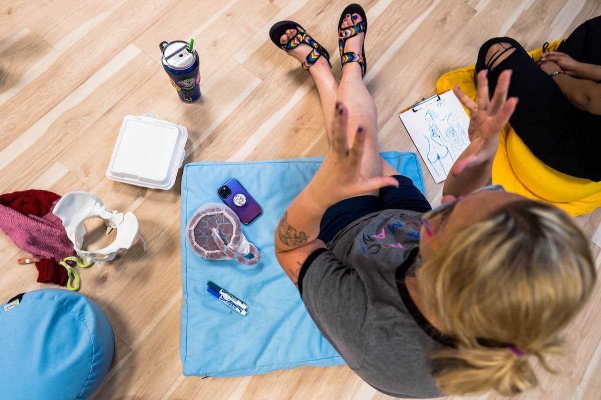 Top view of a person sitting on the floor, gesturing with hands, surrounded by anatomy props, a phone, and drawing materials.