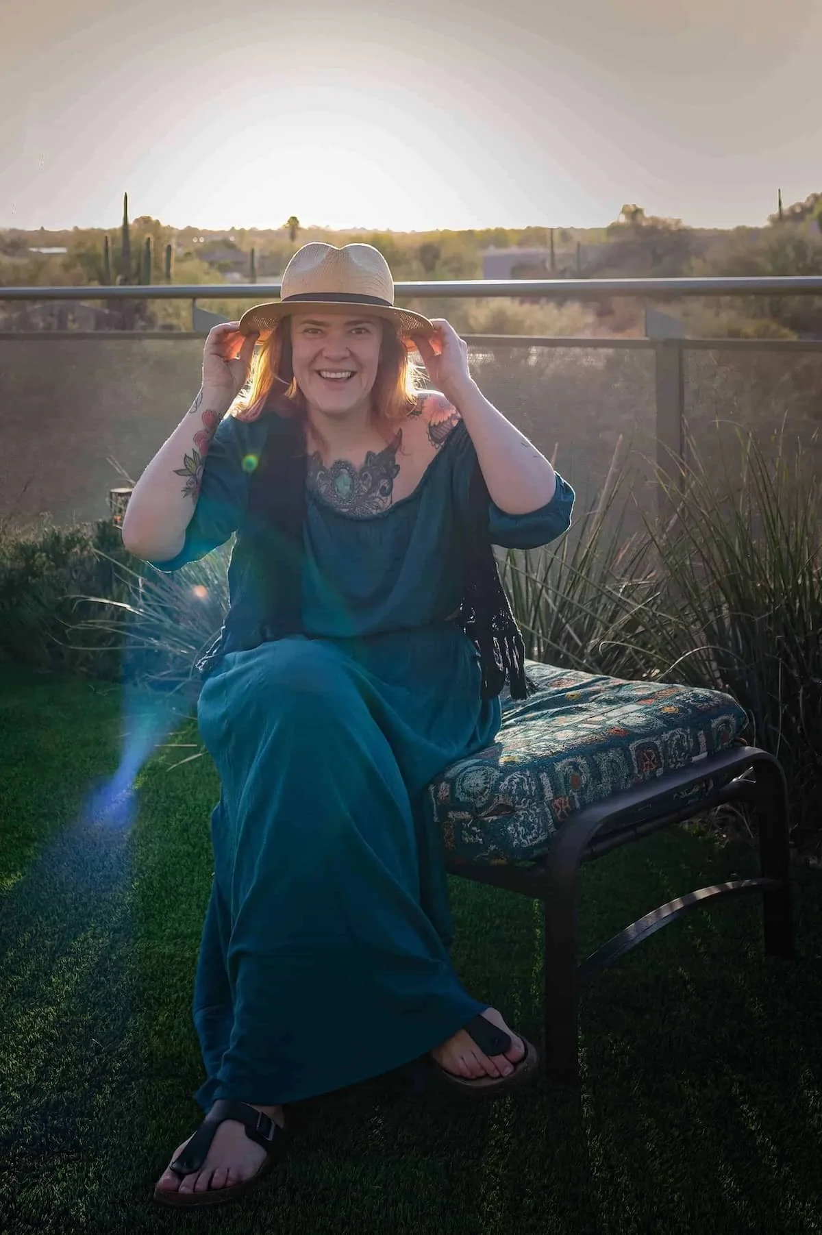 A smiling woman sitting on a patterned outdoor bench, wearing a blue dress, a wide-brimmed hat, and black sandals, with tattooed arms, during sunset with desert landscape in the background.