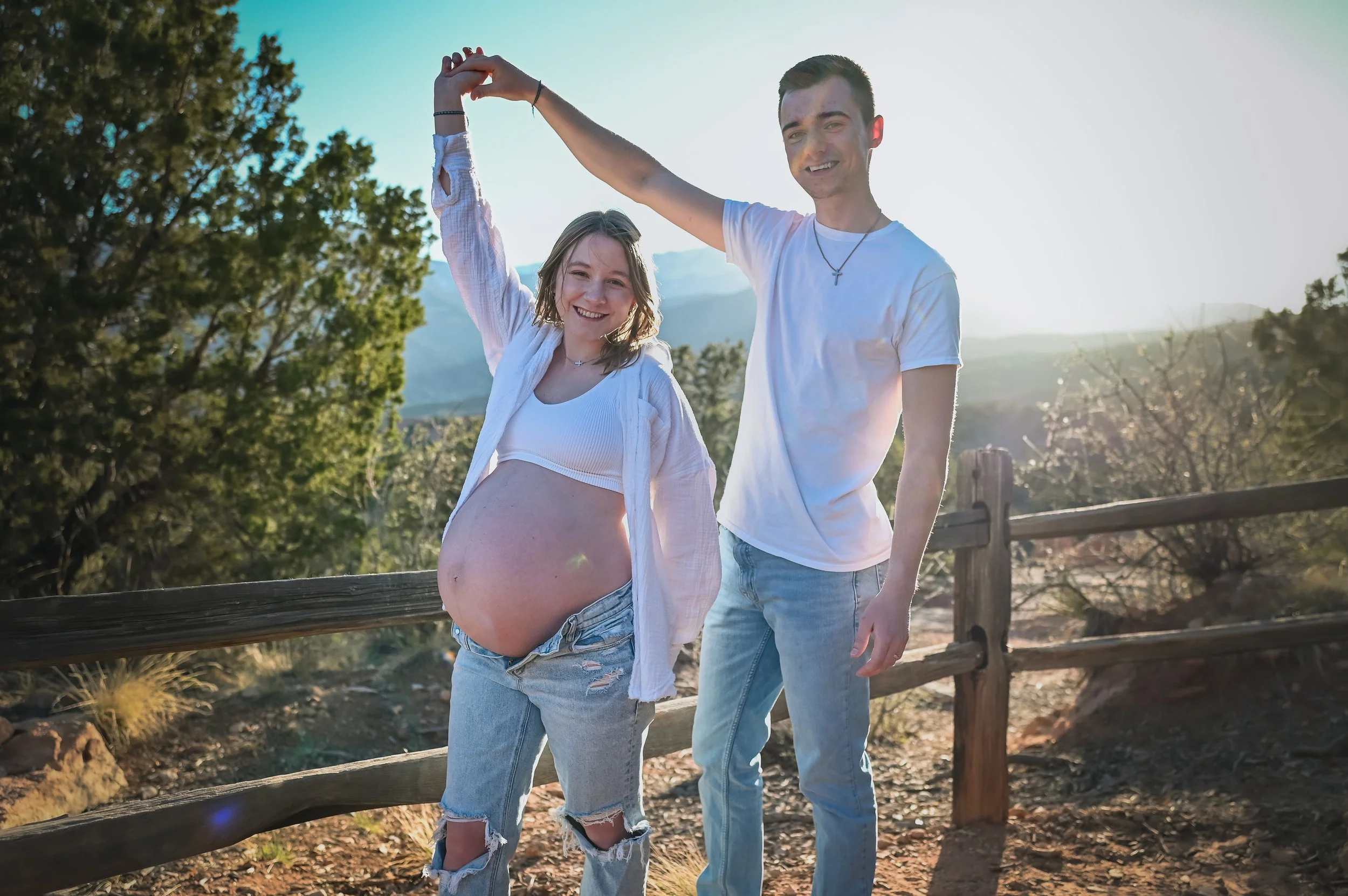 A happy pregnant woman and a young man together outdoors, holding hands raised above their heads. The woman is smiling and wearing a crop top, open shirt, and ripped jeans, with her baby bump visible. The man is smiling and wearing a white t-shirt an