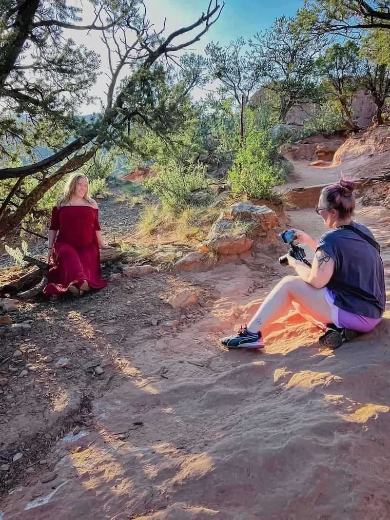 A woman in a red dress poses on a rocky path while another woman kneels nearby, photographing her outdoors.