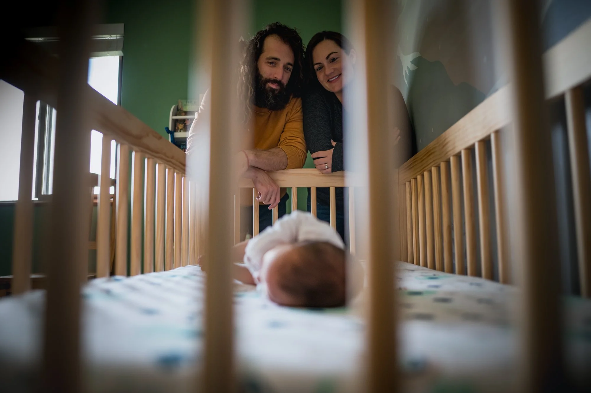 A couple leaning over a crib, smiling at a baby lying on its stomach inside the crib.