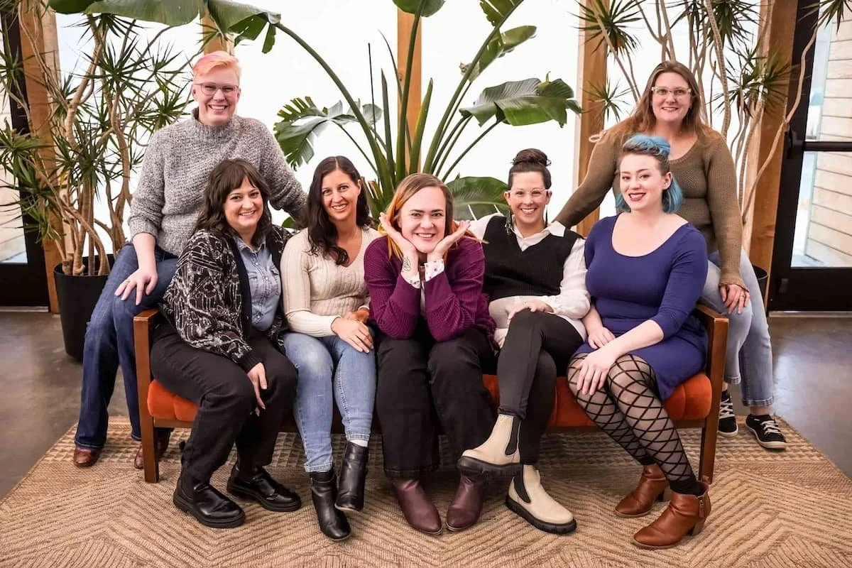A group of seven women sitting and standing around a wooden bench in a room with large plants and natural light, smiling at the camera.