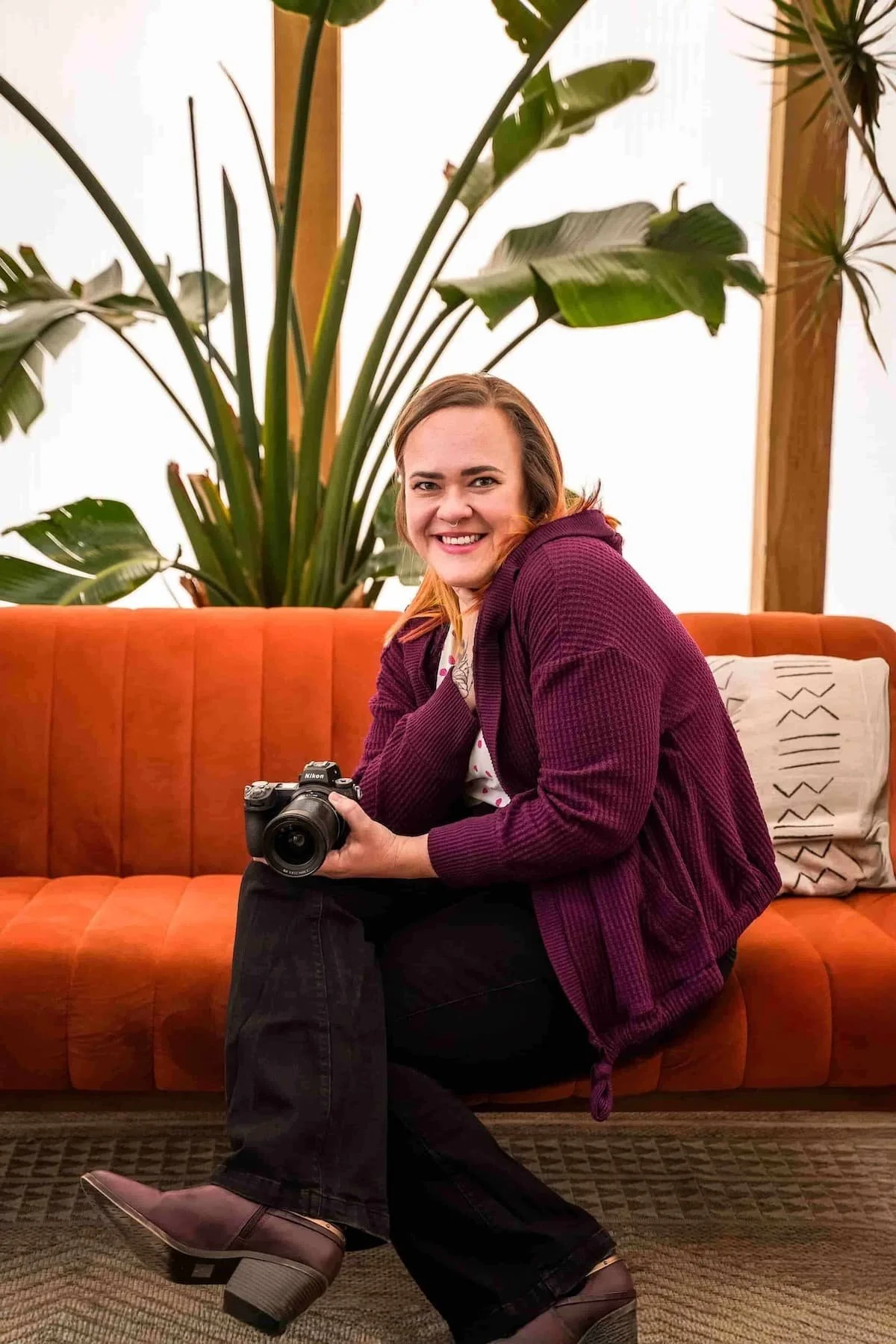 A woman with light skin and red hair smiling while sitting on a burnt orange couch, holding a camera in her right hand. She is wearing a purple sweater, black pants, and brown ankle boots. Behind her is a large green plant and a white pillow with black geometric patterns.