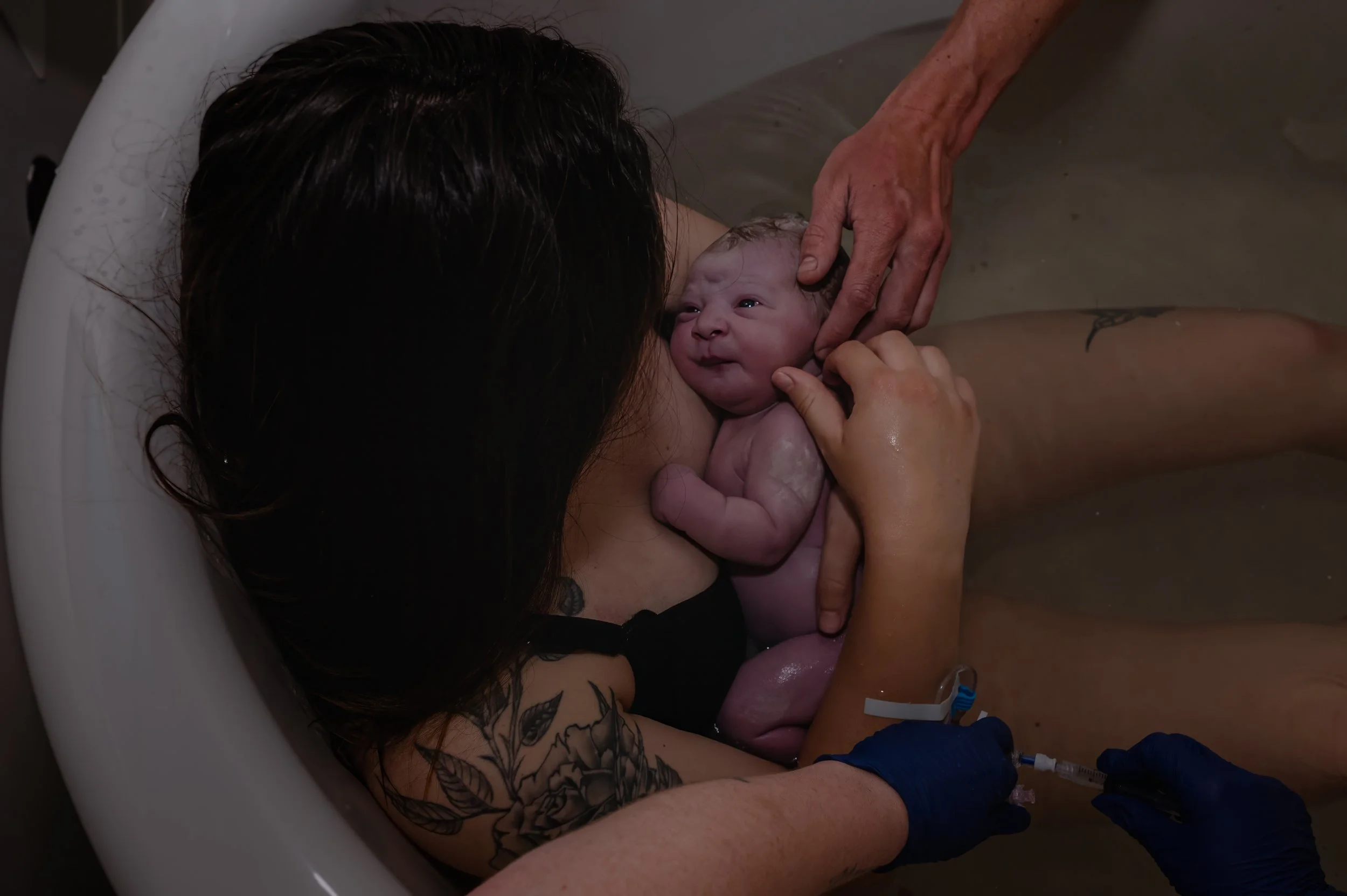 A woman giving birth in a birthing tub, holding her newborn baby close to her chest while a person with gloves administers a medical injection to her leg.