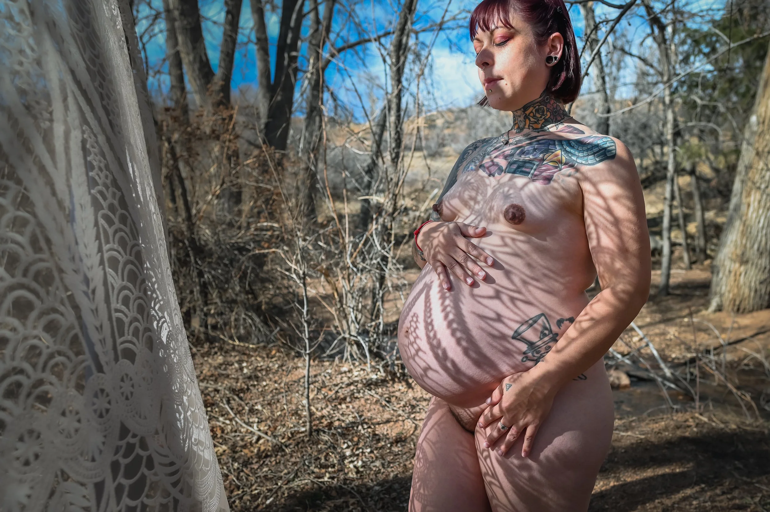 A nude woman standing outdoors in a forest, with shadows of leaves creating patterns on her body and face.