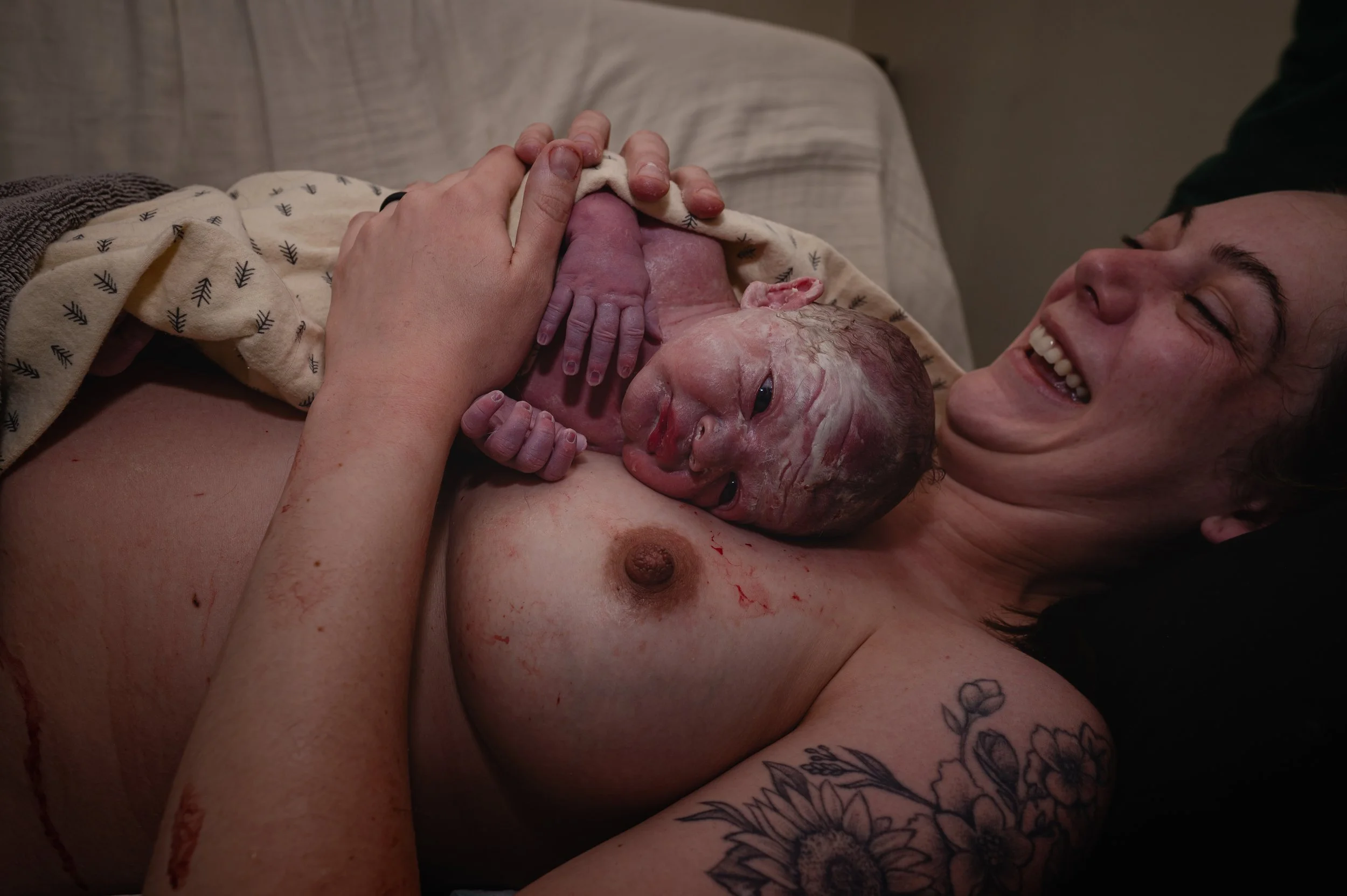 A woman with tattoos on her arm, holding a newborn baby on her chest, is smiling. The scene appears to be a delivery in a hospital or home setting.