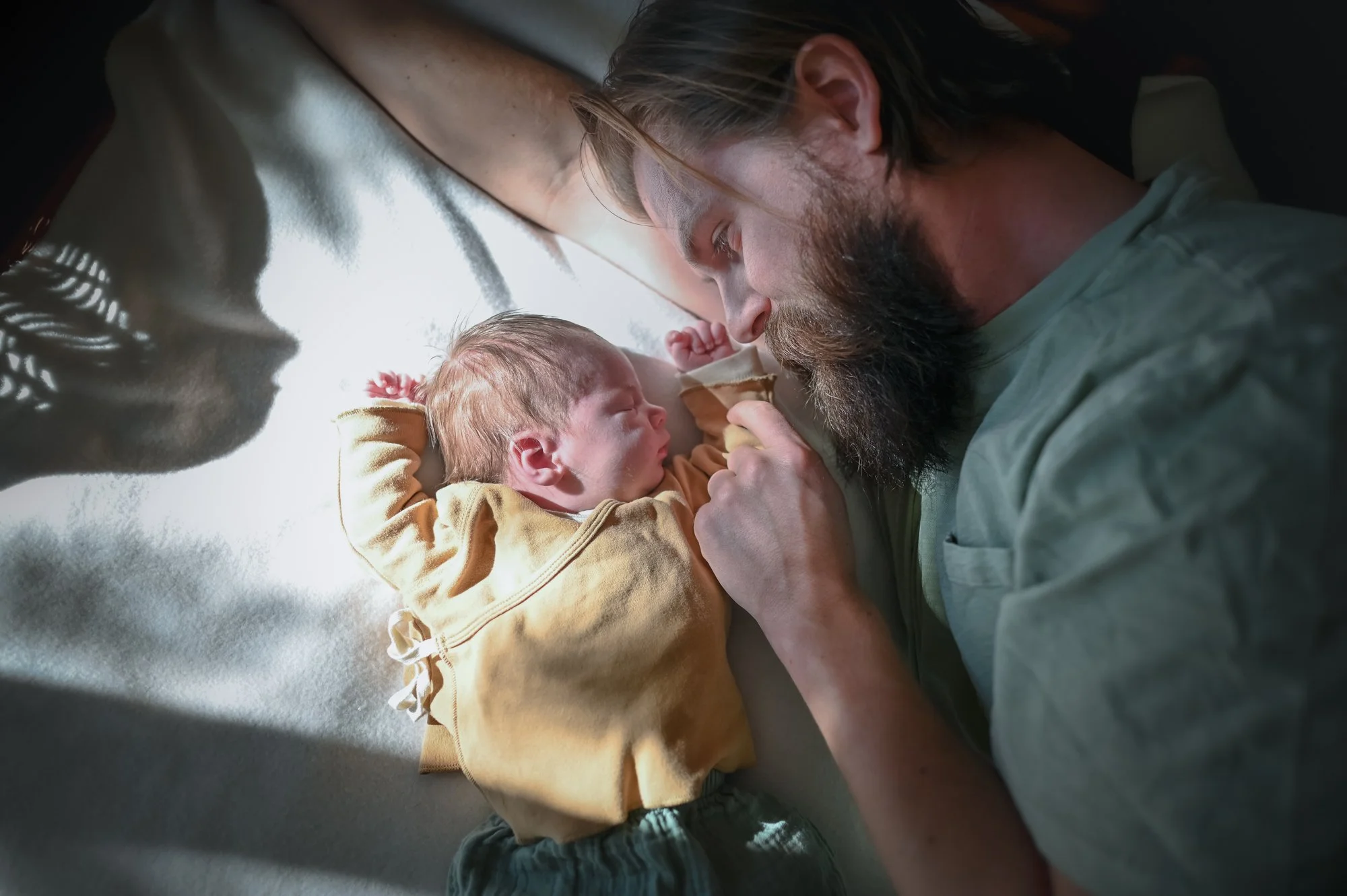 A father and newborn baby lying on a bed, facing each other, with natural light casting shadows. The father has a beard and long hair, and is gently touching the baby's hand.