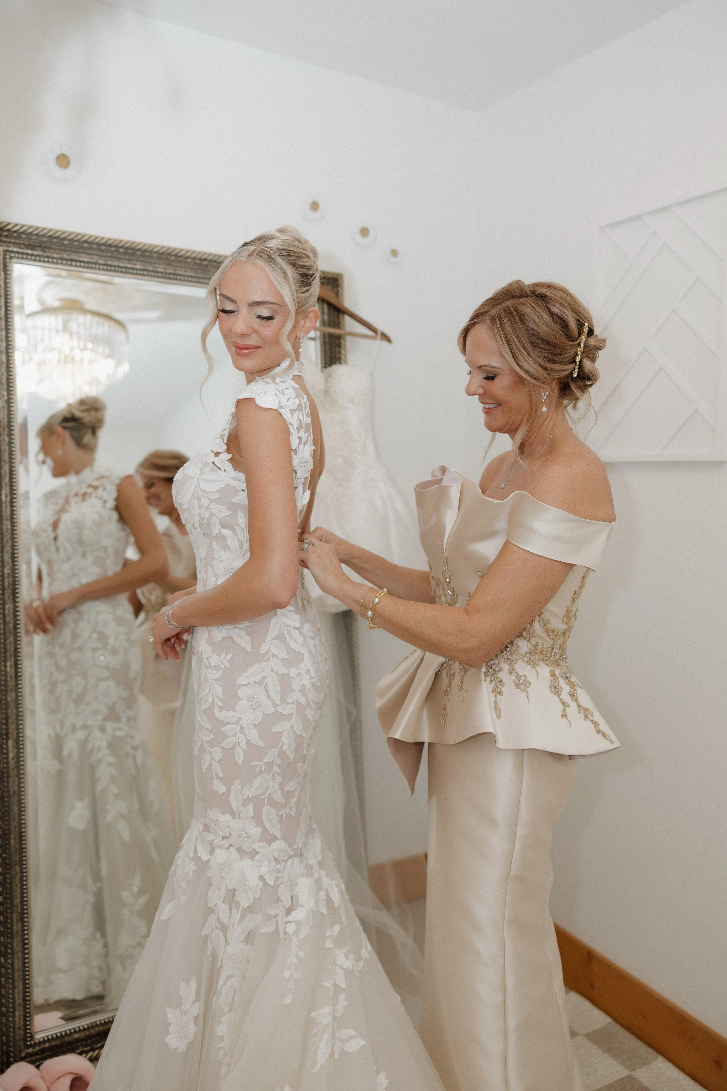 A bride and her mother share a special moment as the mother helps fasten the back of the bride's wedding dress in a room with a large mirror and elegant chandelier.