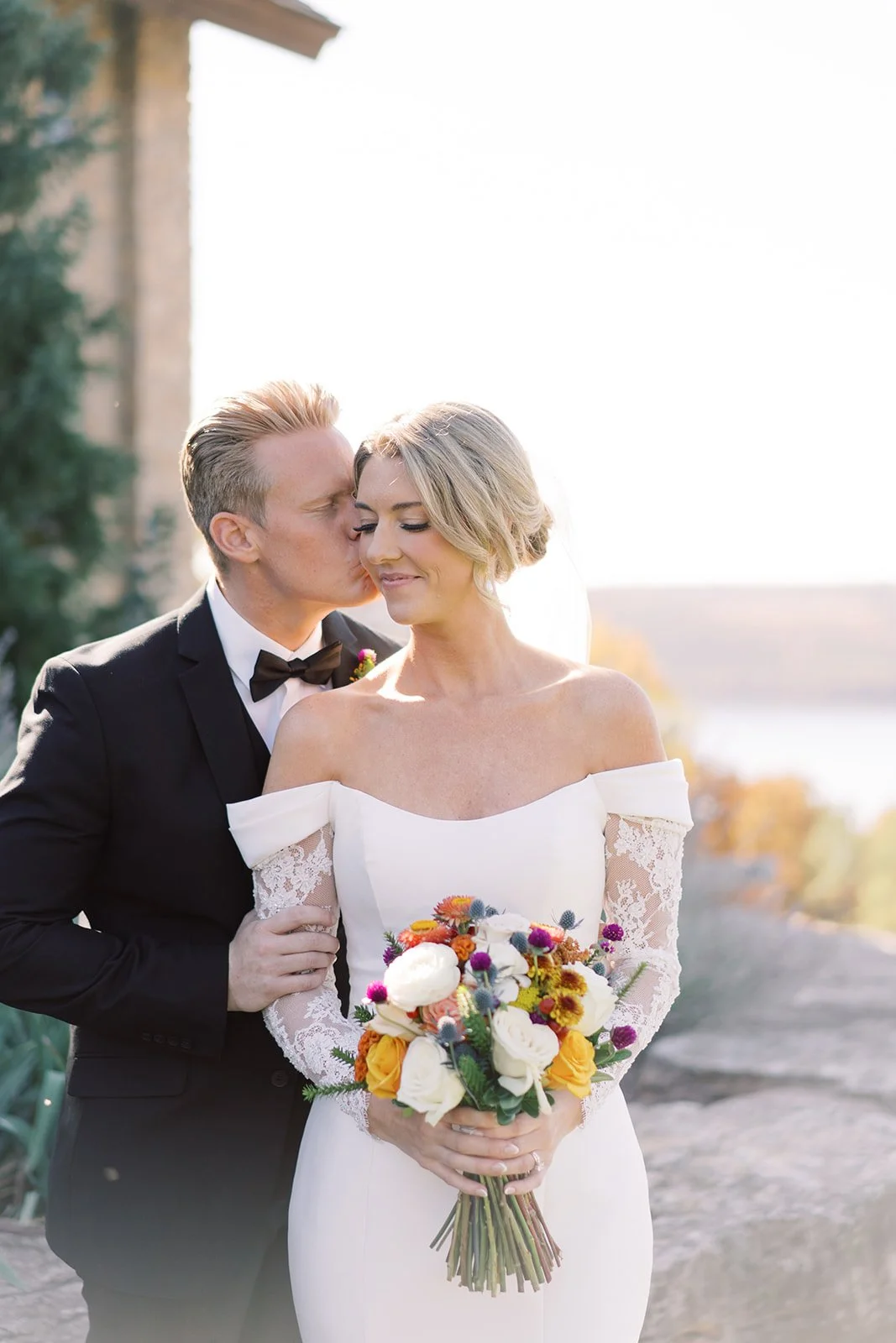 A bride and groom stand outdoors during their wedding, with the groom kissing the bride on her forehead. The bride is holding a colorful bouquet of flowers and is wearing an off-the-shoulder wedding dress. The groom is dressed in a black tuxedo with a bow tie.