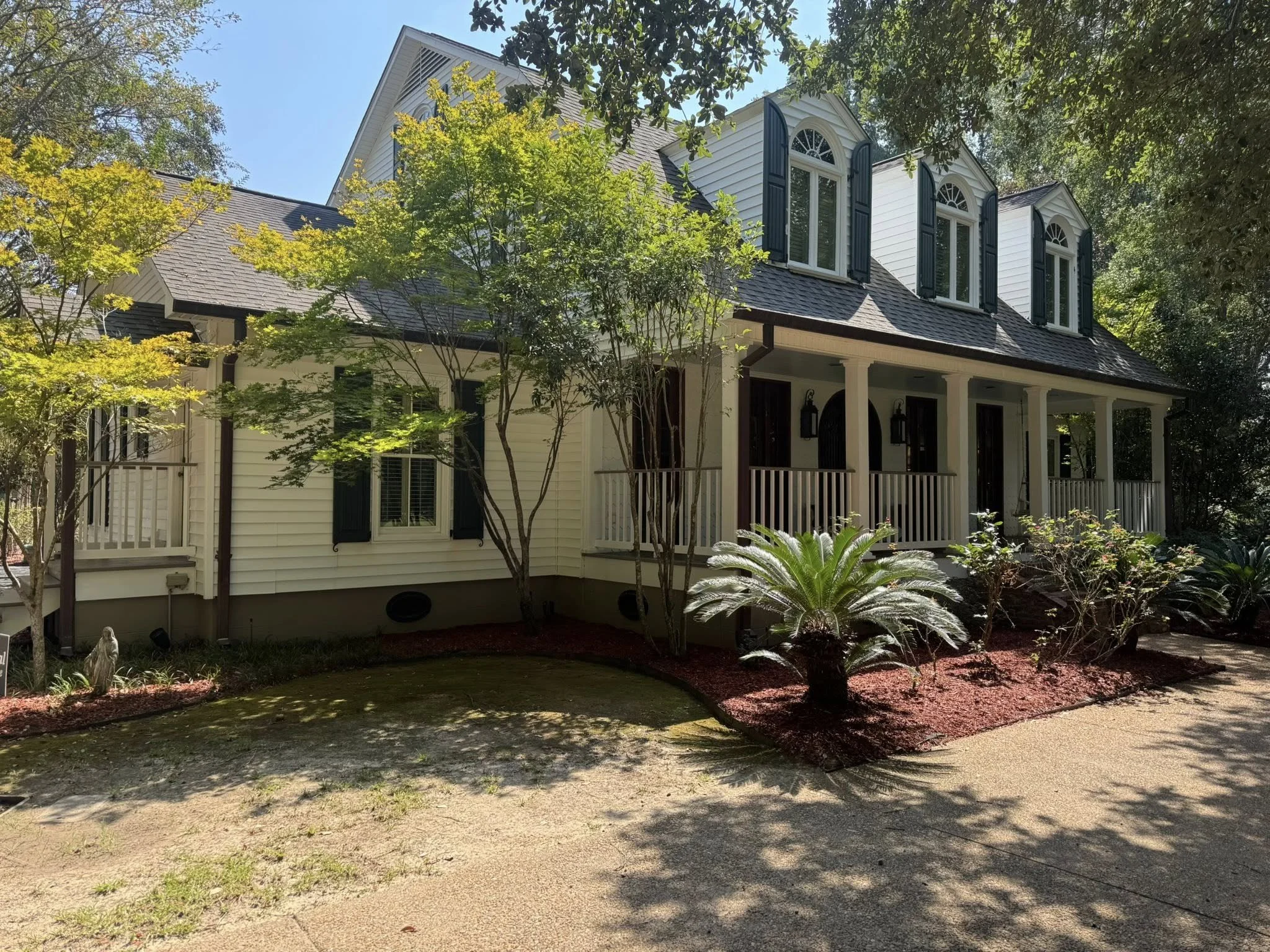 A house with white siding, black shutters, and shingled roof, surrounded by trees and a landscaped yard with bushes and mulch, under a blue sky.